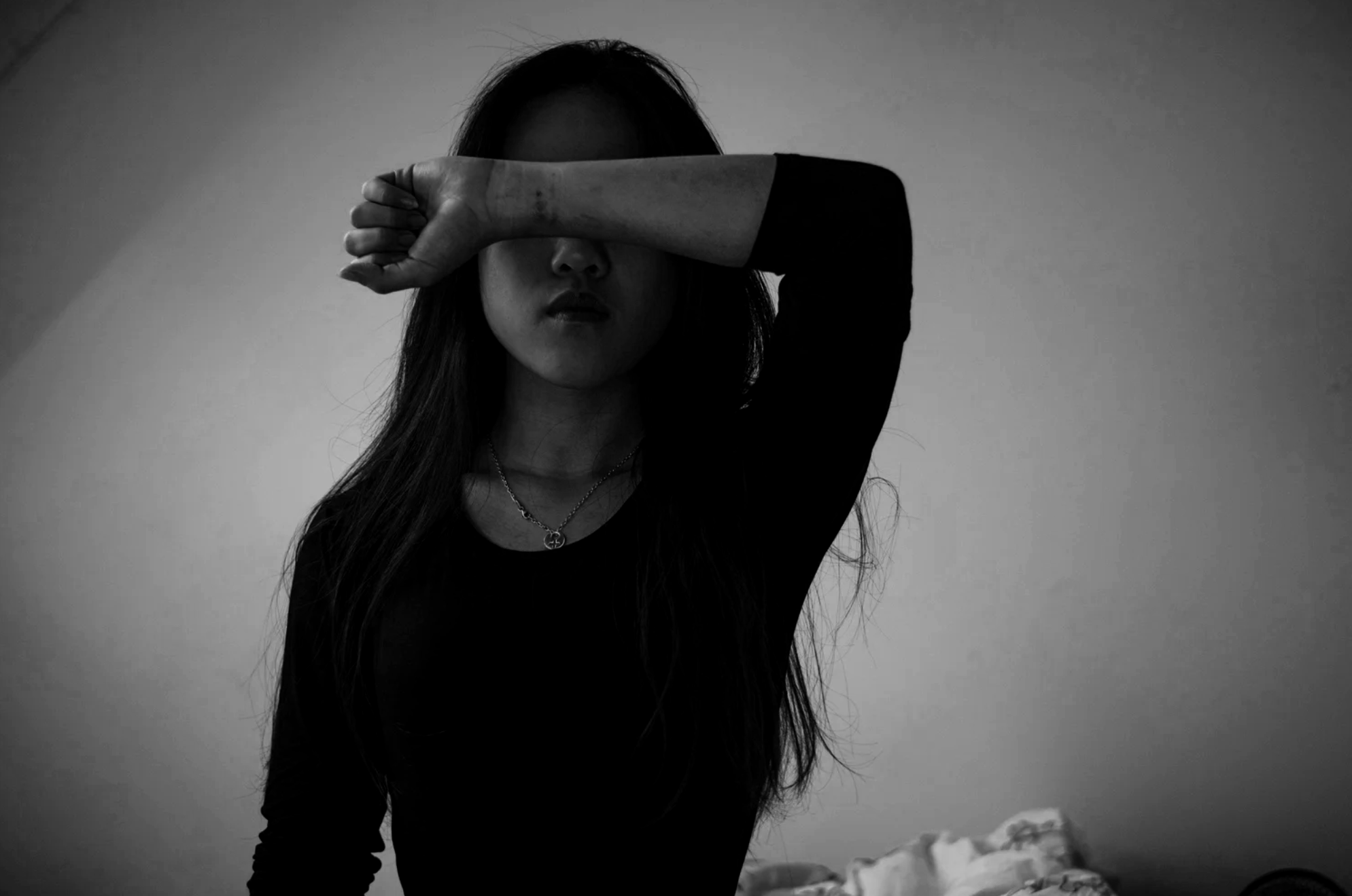 A woman with long hair is partially covering her face with her arm. The photo is in black and white and she is wearing a dark top and a necklace.