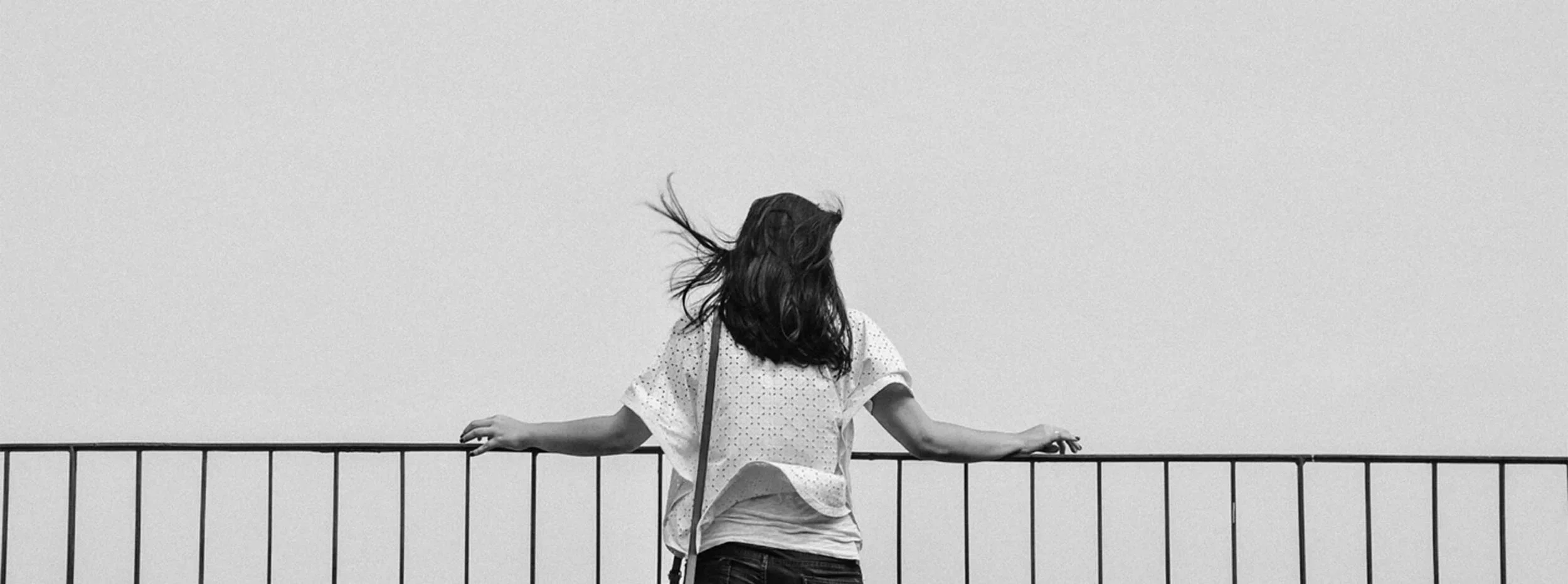 A woman with long hair facing away, standing behind a metal railing, with her hair blowing in the wind against a plain sky.