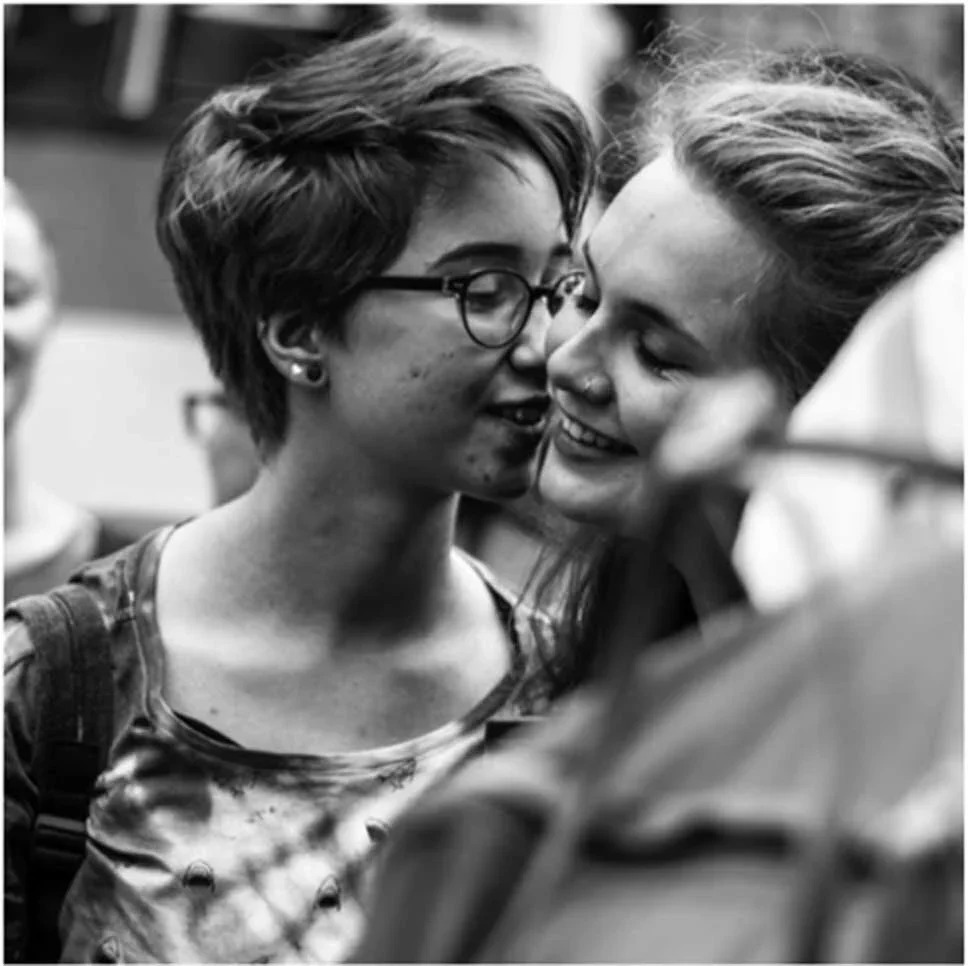 Two young women sharing a close moment, smiling and leaning in with noses touching, in a social setting.