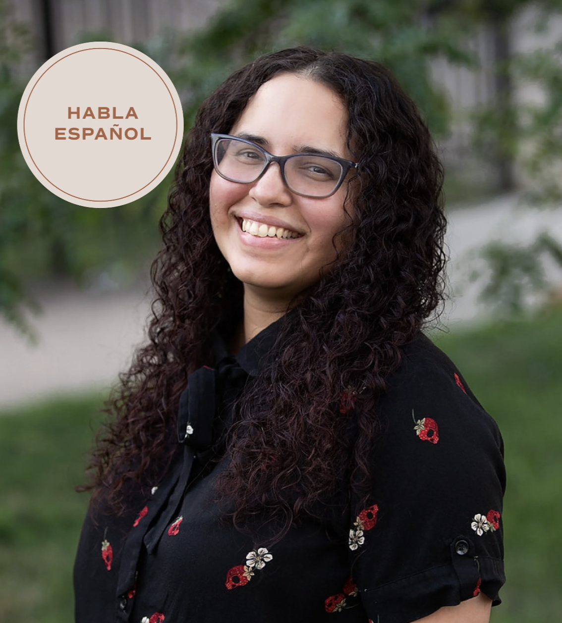 A smiling woman with curly dark hair and glasses outdoors, wearing a black shirt with red and white floral embroidered designs, with trees and greenery in the background, and a circular label that says 'Habla Español'.
