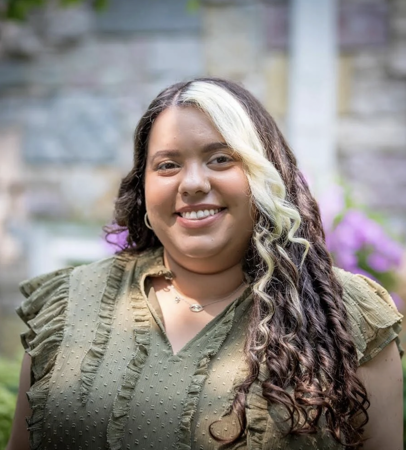 A woman with long, curly hair, some of which is dyed blonde, smiling in front of a blurred outdoor background with purple flowers.