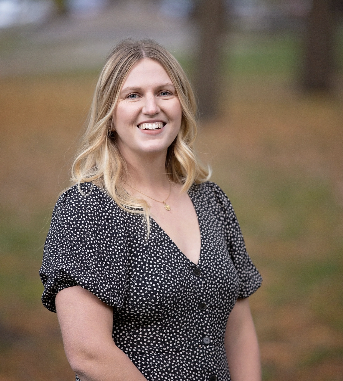 A smiling woman with blonde wavy hair wearing a black dress with white polka dots, standing outdoors in a park-like setting.