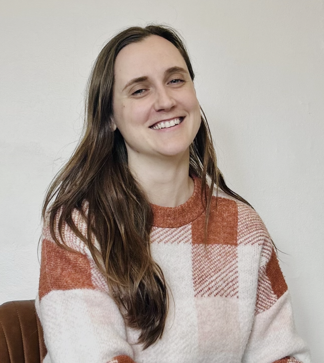 A smiling woman with long brown hair wearing a red and white plaid sweater, sitting against a plain white wall.