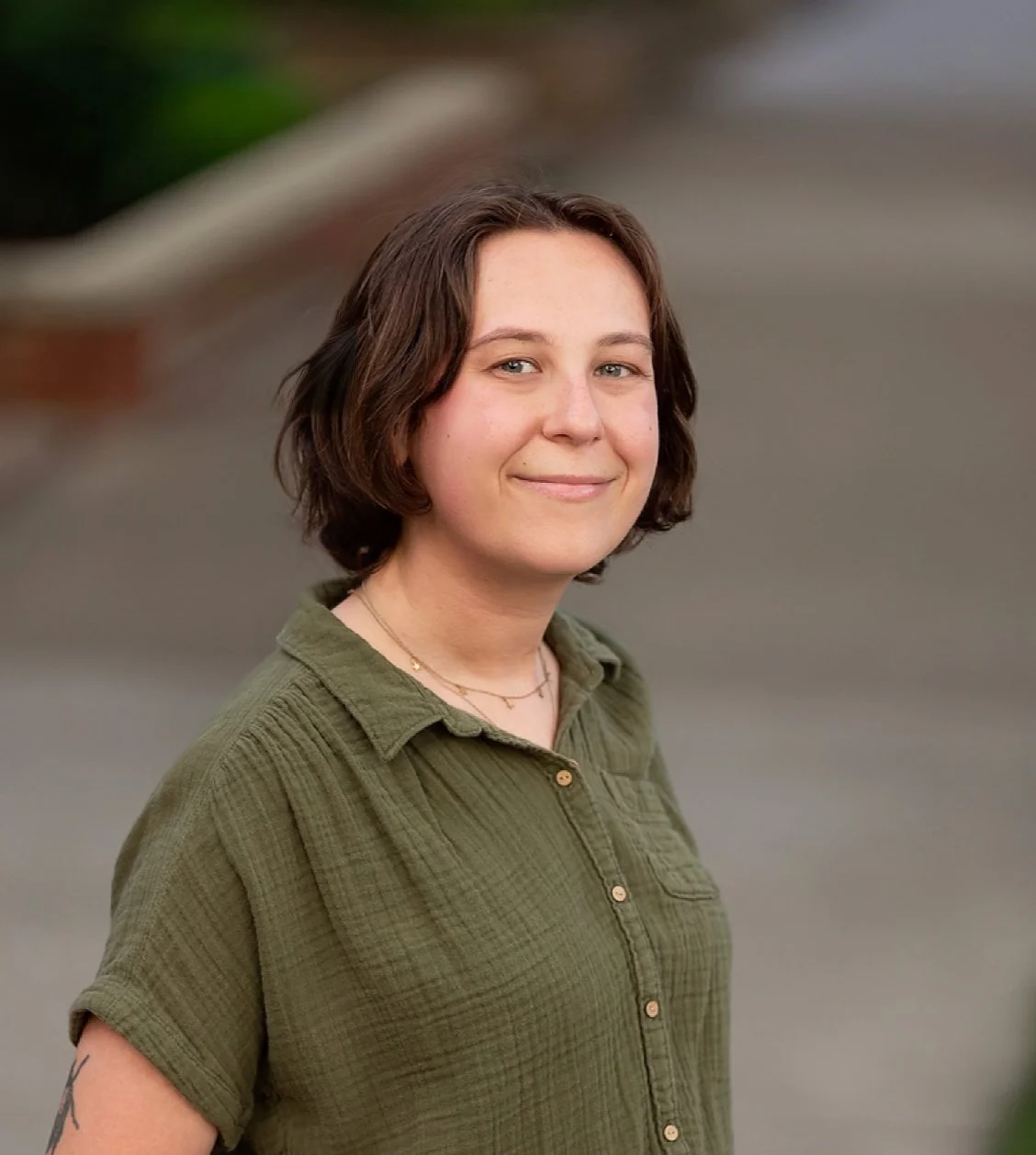 A young woman with short brown hair and light skin smiling outdoors, wearing a green button-up shirt.