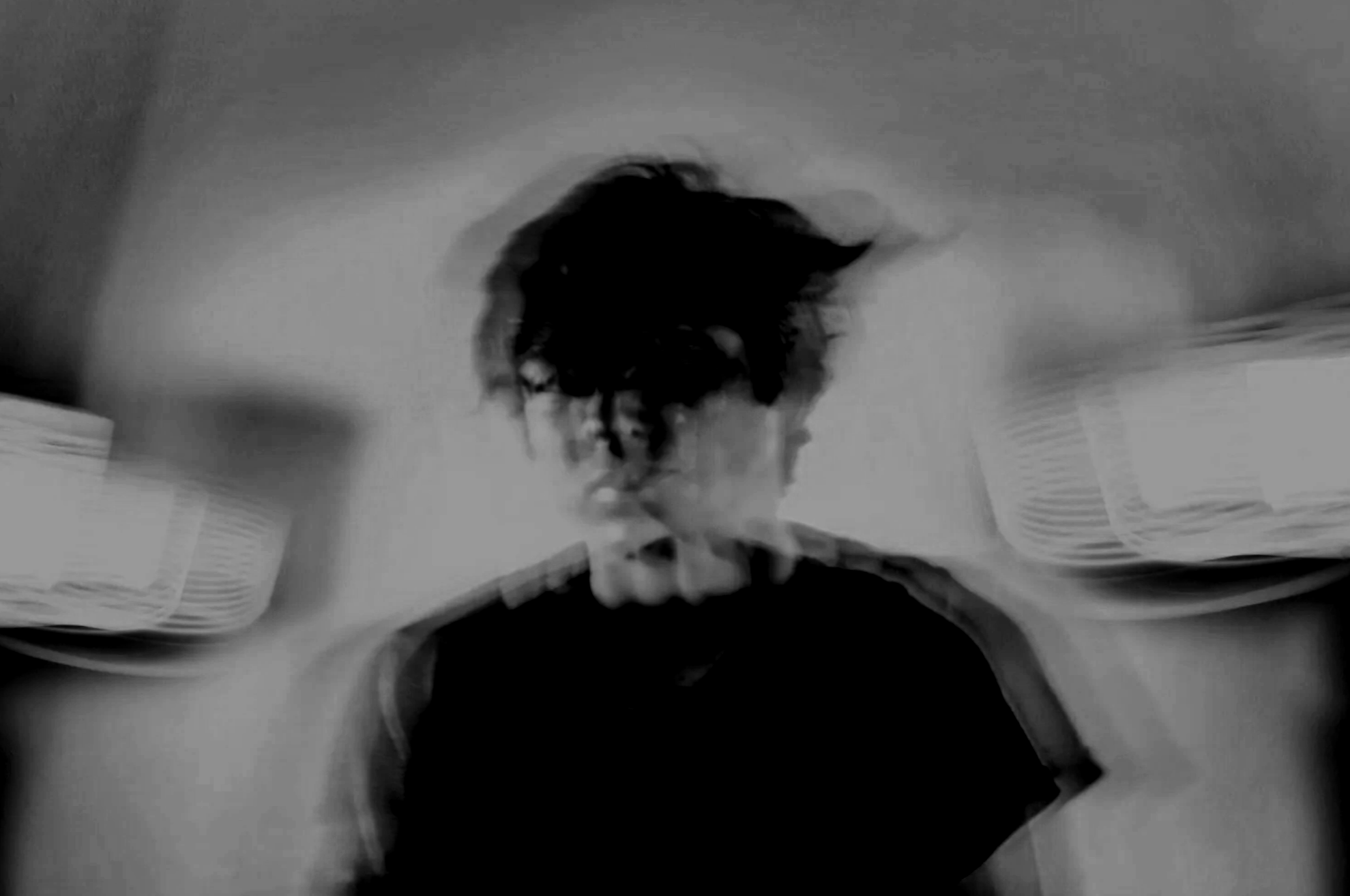 Black and white distorted image of a person with dark, messy hair, standing in a room with shelves or storage bins on the walls.