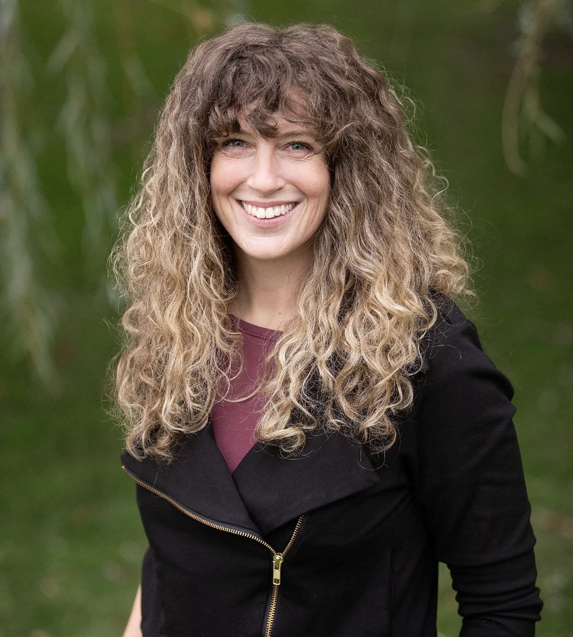 Smiling woman with long, curly hair, wearing a dark jacket and maroon top, outdoors with green foliage in the background.