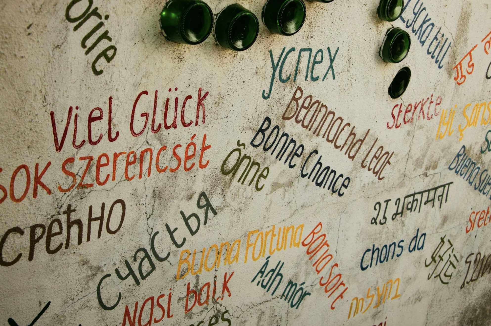 A wall with colorful handwritten words in various languages and scripts, along with several green bottle caps attached to the wall.