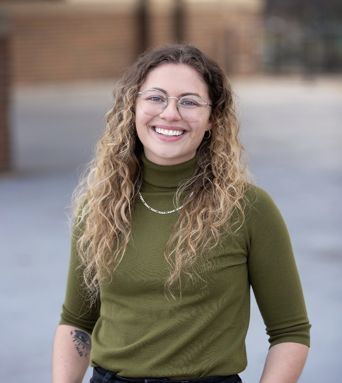 A young woman with curly blonde hair, wearing glasses, a green turtleneck, and jewelry, smiling outdoors.