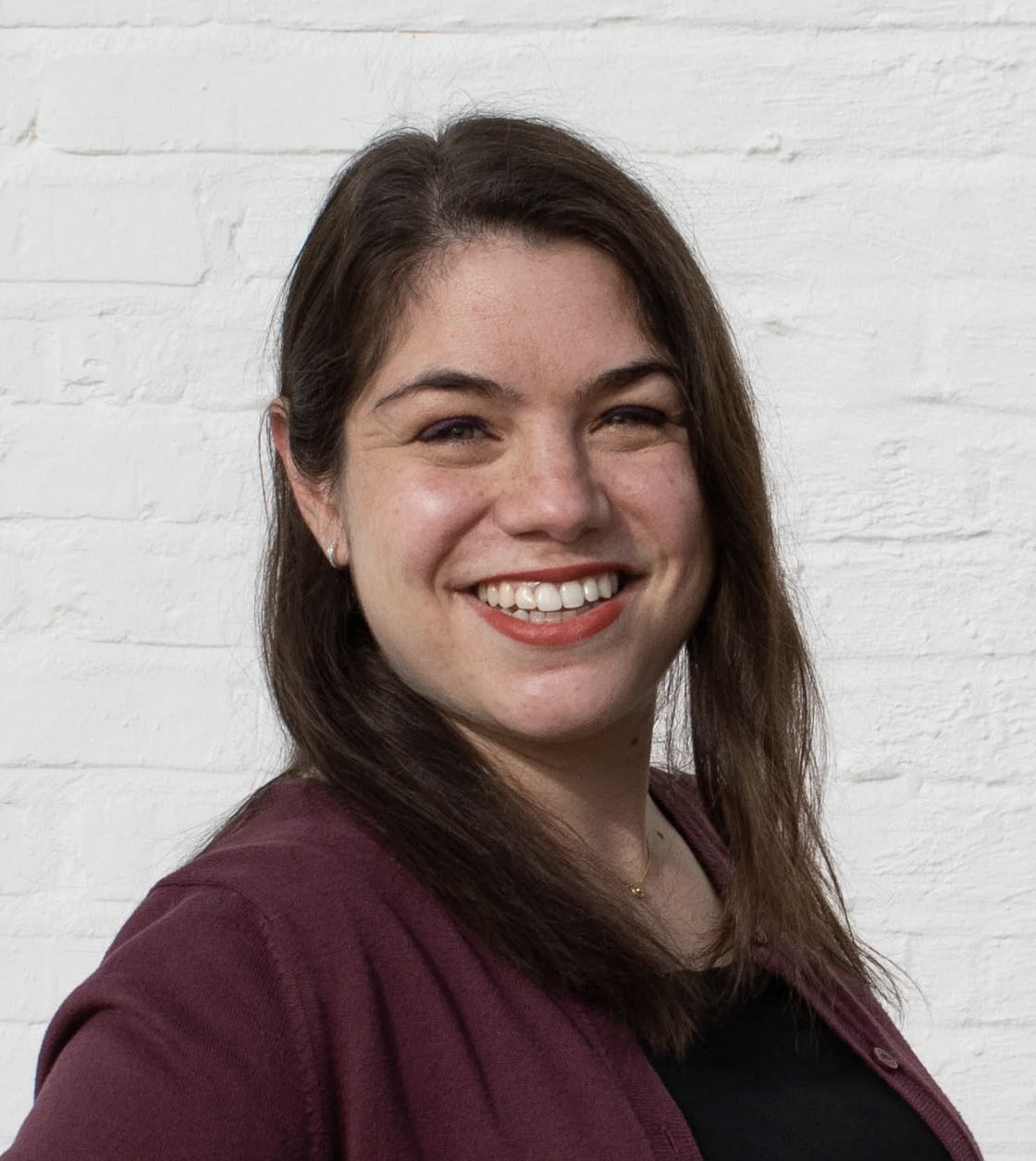 A woman with long brown hair, smiling, wearing a maroon jacket and a black top, standing against a white brick wall.