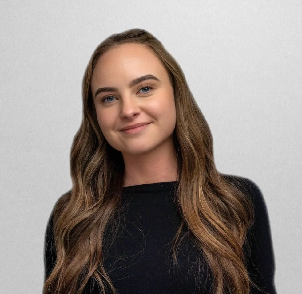 A young woman with long wavy brown hair, wearing black glasses, a black T-shirt, and standing against a plain white background.