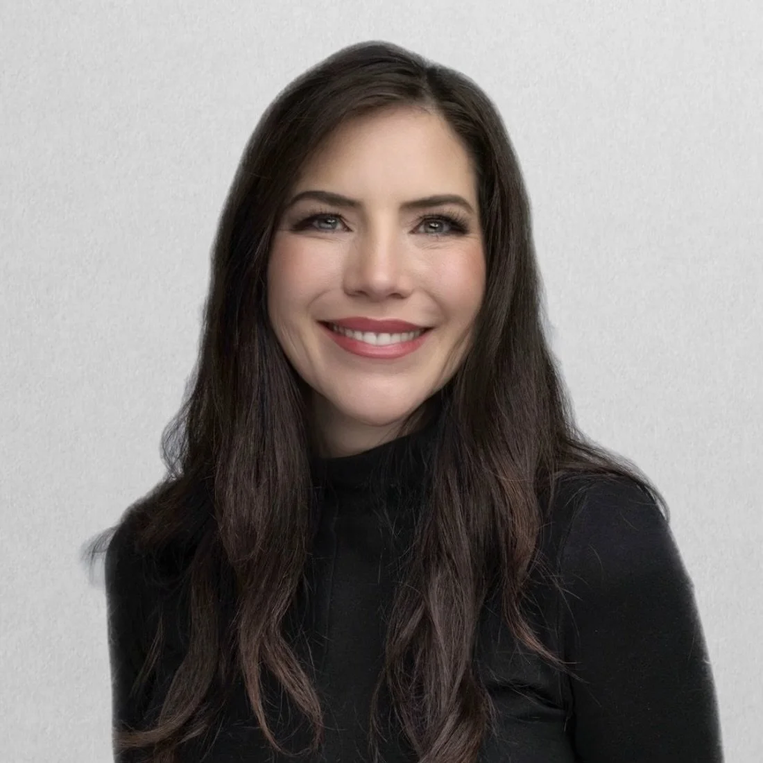 A woman with long dark brown hair, light makeup, and a big smile wearing a black top, standing against a plain light-colored background.