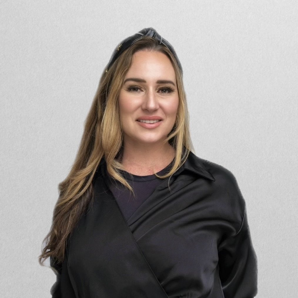 A woman with long, wavy blonde hair and neutral makeup, wearing a black top, posing against a dark background.