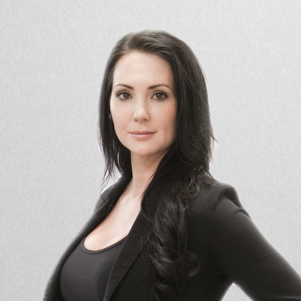 A woman with dark, wavy hair wearing a white long-sleeve top and black pants with a black belt featuring a gold GG buckle, leaning against a white counter in front of a dark blue wall.
