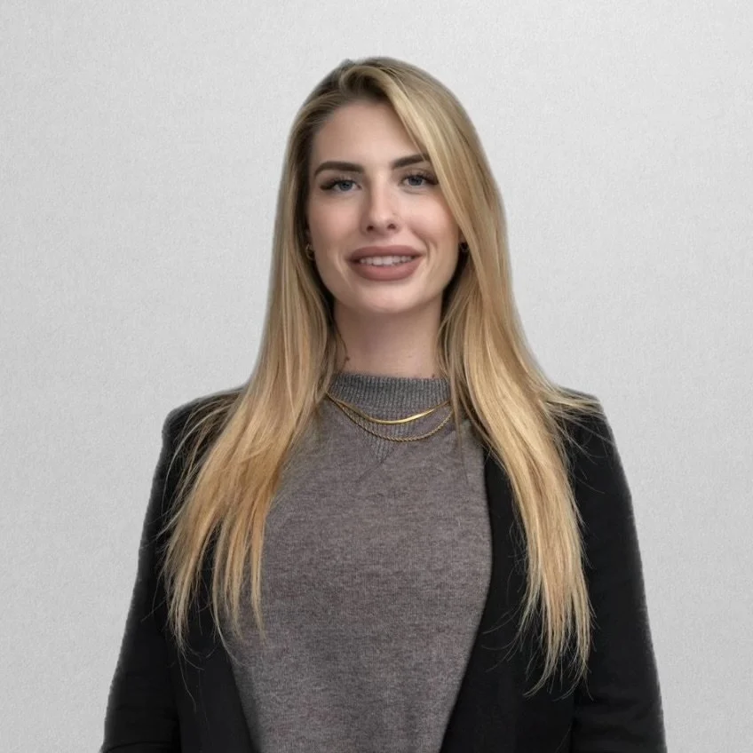 A woman with long blonde hair, wearing a gray top and black blazer, standing against a plain light-colored background.