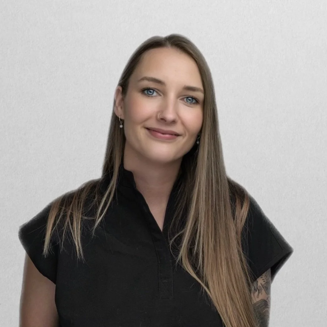 A young woman with long blonde hair, blue eyes, and makeup, smiling while wearing a black blazer and a white shirt.