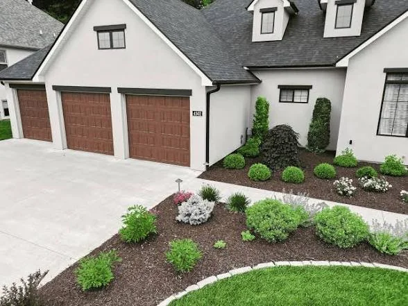 Front yard landscaping with small bushes, shrubs, and flowers in mulch beds in front of a white house with brown garage doors.