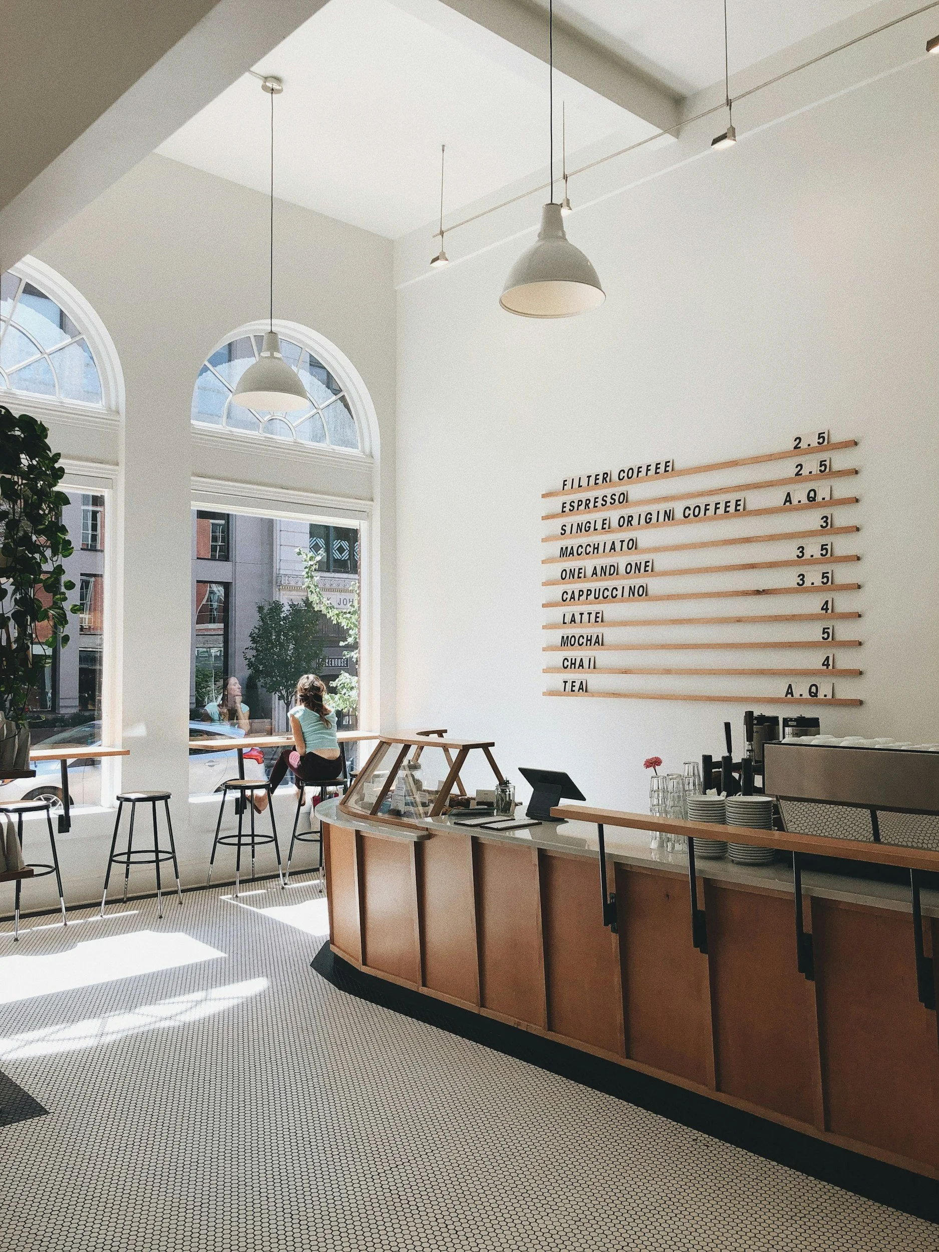 Interior of a modern coffee shop with a white wall, wooden counter, and large arched windows letting in natural light. There are a few customers sitting by the windows, and a menu board on the wall listing various coffee drinks with prices.
