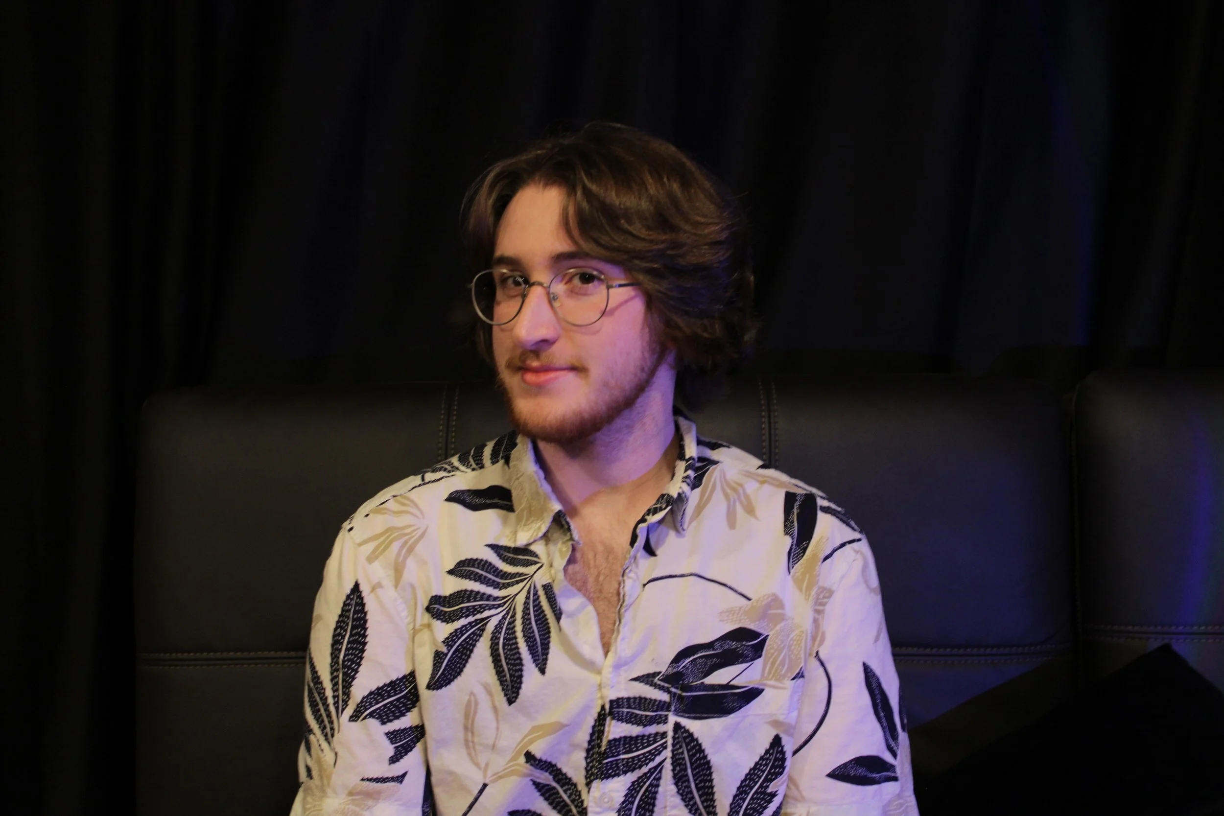 A young man with wavy brown hair, glasses, and a beard, wearing a floral-patterned shirt, sitting on a black leather couch with a dark curtain background.