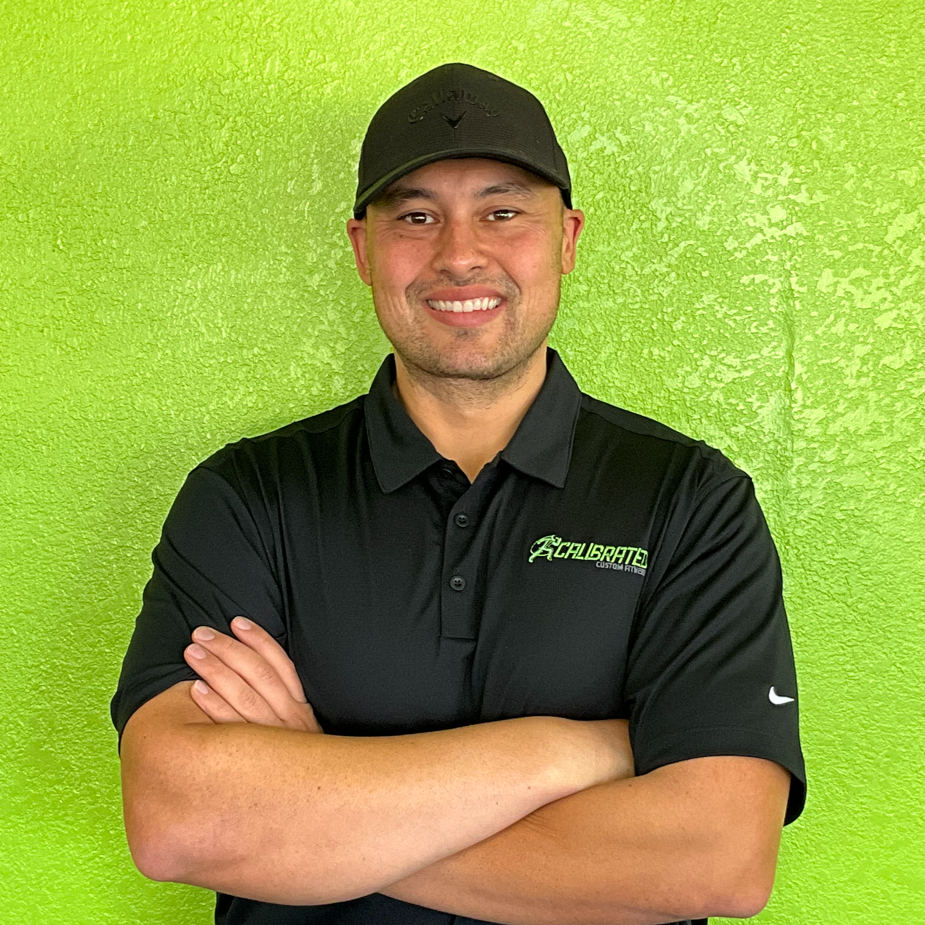 A man with light skin, wearing a black baseball cap and black polo shirt, stands in front of a neon green wall, smiling with arms crossed.