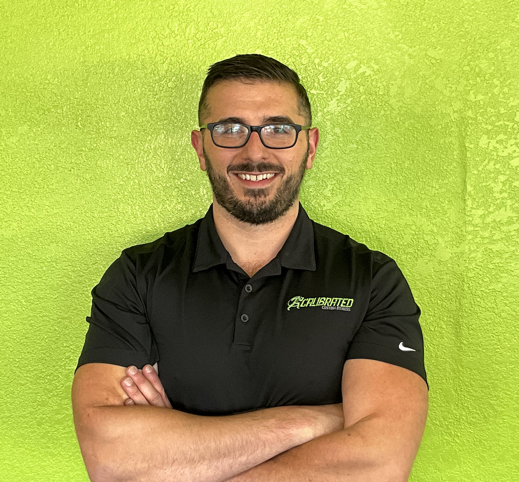 Man smiling with arms crossed, wearing glasses and a black polo shirt with logo, in front of a bright green textured wall.
