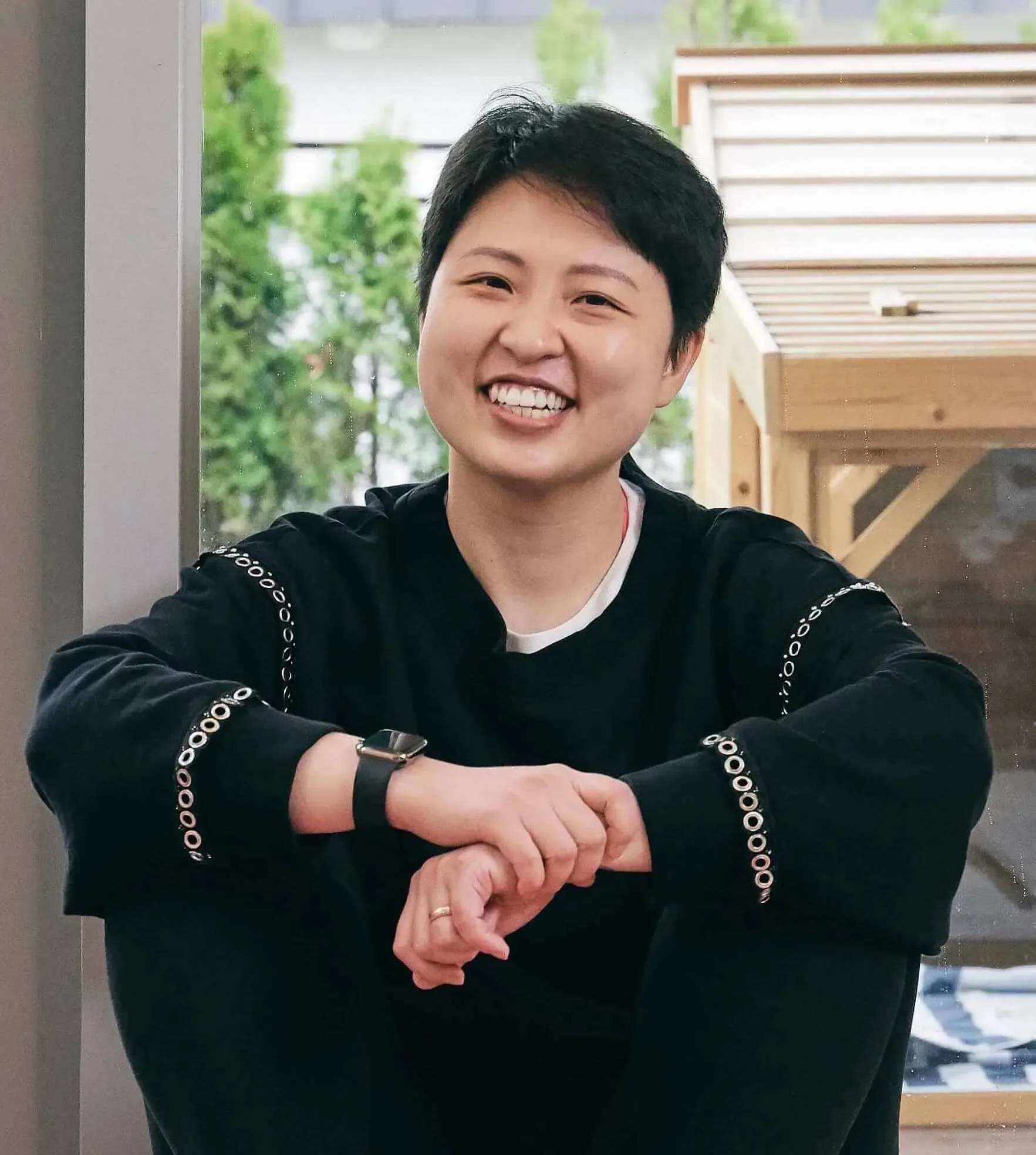 A woman with short black hair, smiling, wearing a black long-sleeve shirt with circular patterns on the sleeves, sitting with her arms resting on her knee, in what appears to be a modern indoor setting with a large window and green outdoor foliage in the background.