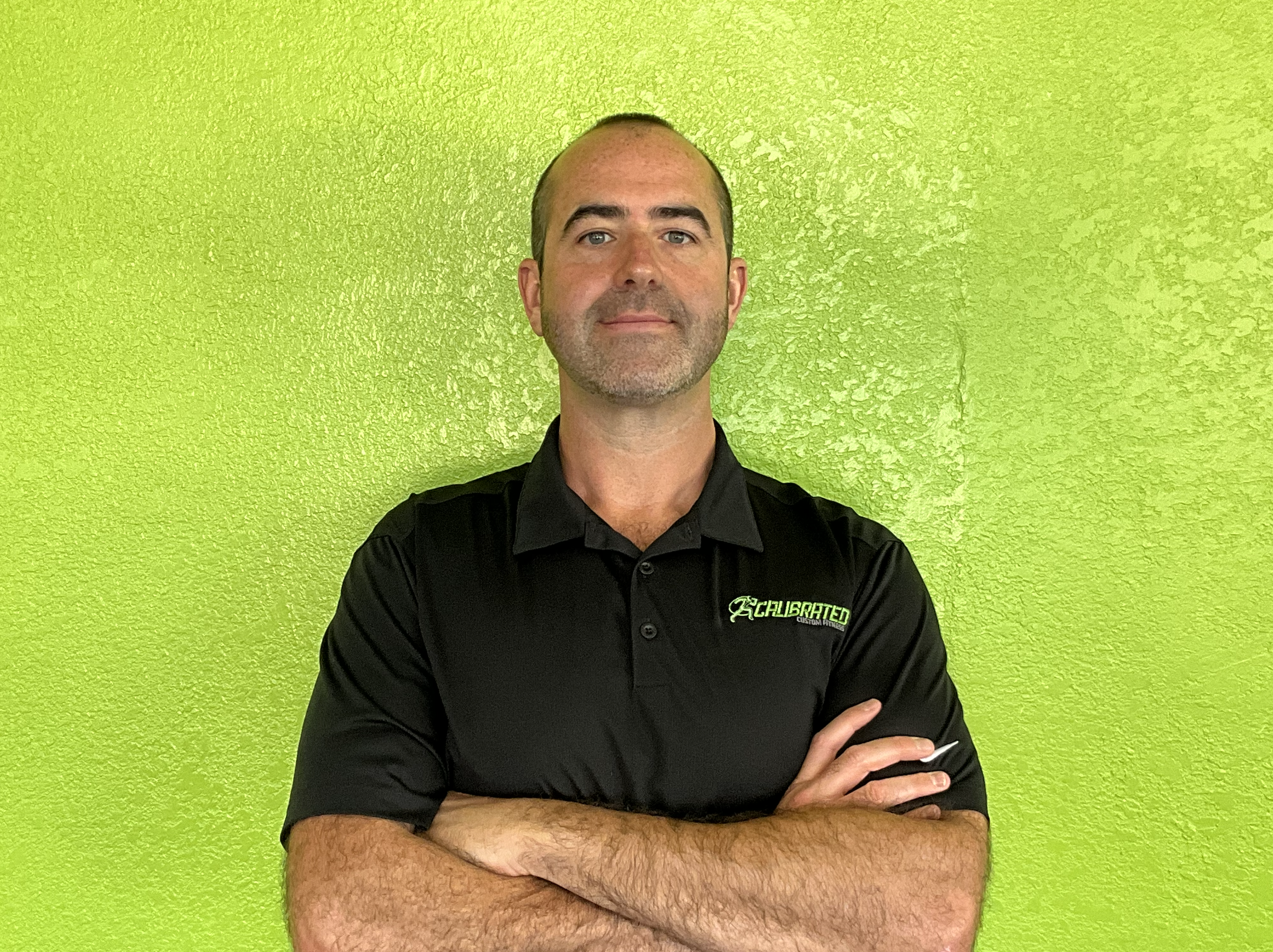 A man with arms crossed, standing in front of a bright green wall, wearing a black polo shirt with a logo on the left side.