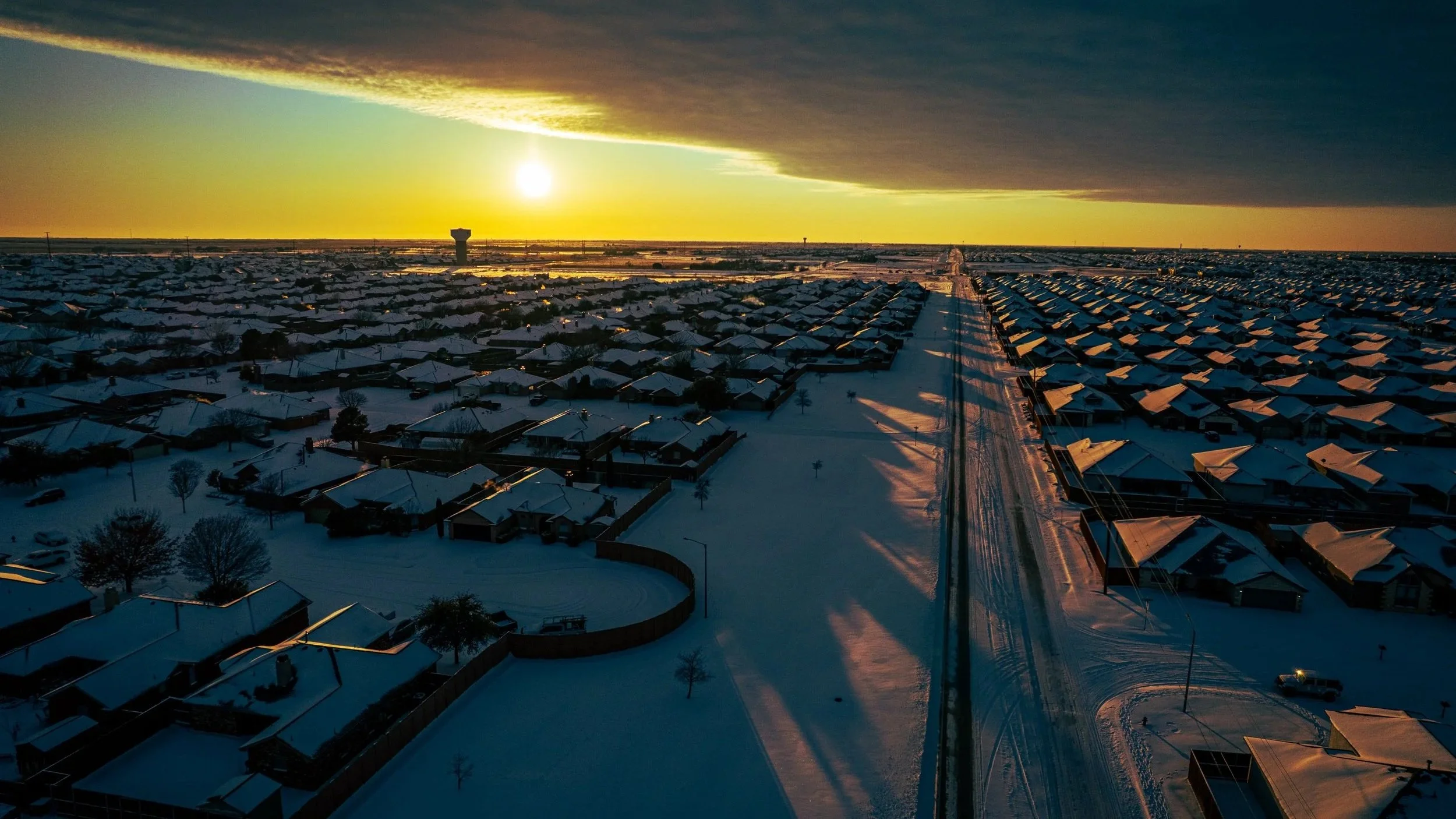 Snow covers a suburban neighborhood as a colorful sunset illuminates the sky, creating a shadow and colorful effect on the snow below.