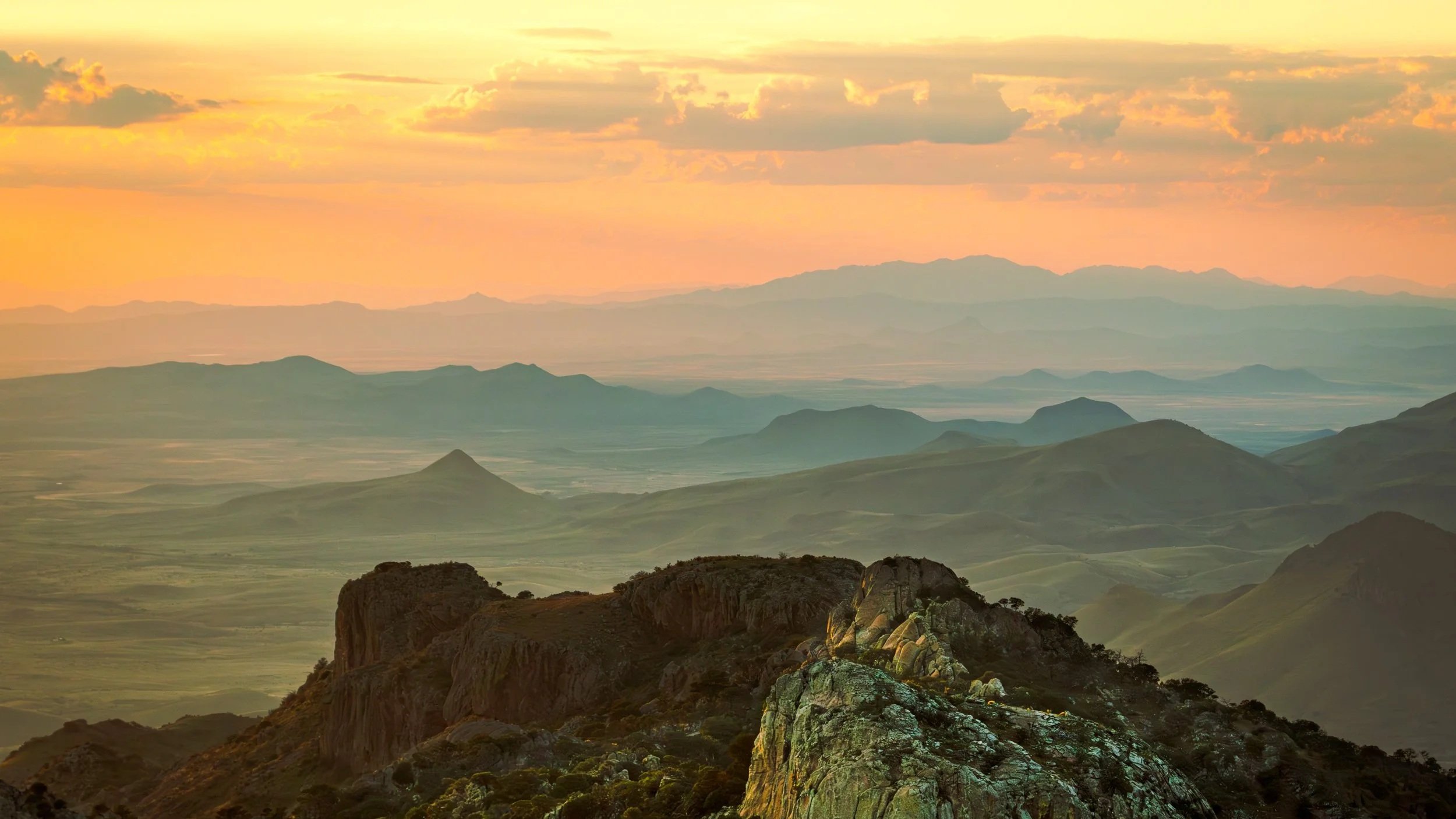 Scenic landscape of vast layered rugged mountains at sunset with fog, clouds, and warm pastel colors in the sky.