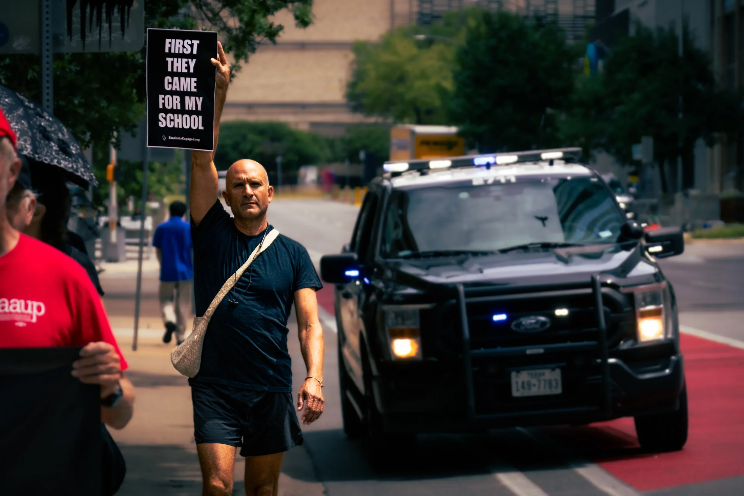 A protestor marching holds a sign reading "First they came for my school" as a police cruiser ominously drives on the road behind him.