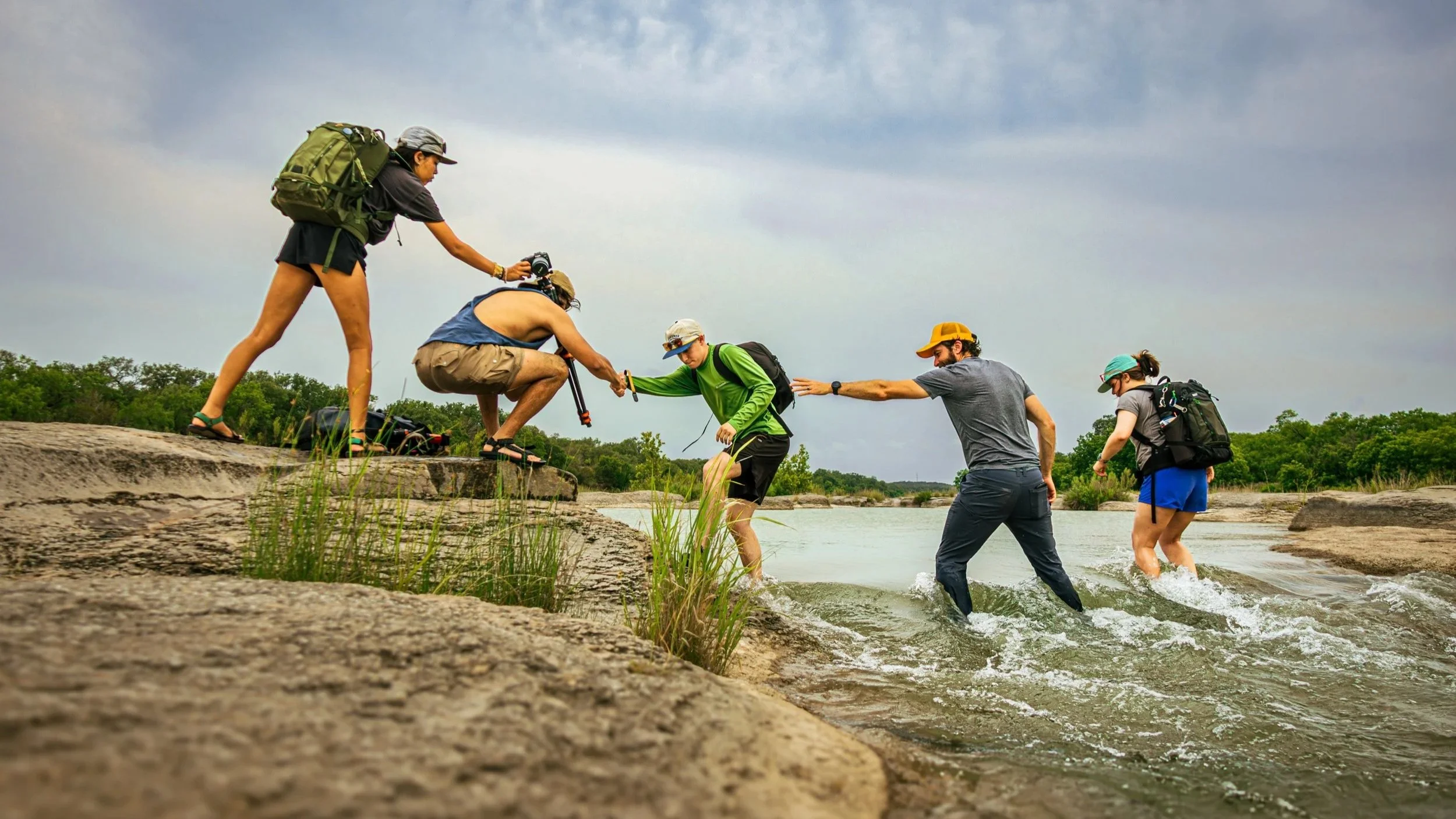 Five hikers with backpacks and hats are helping each other across a shallow creek with rapid waters on a cloudy day. They are all holding on to each other and interconnected, creating a visual reminiscent of a paper doll chain.