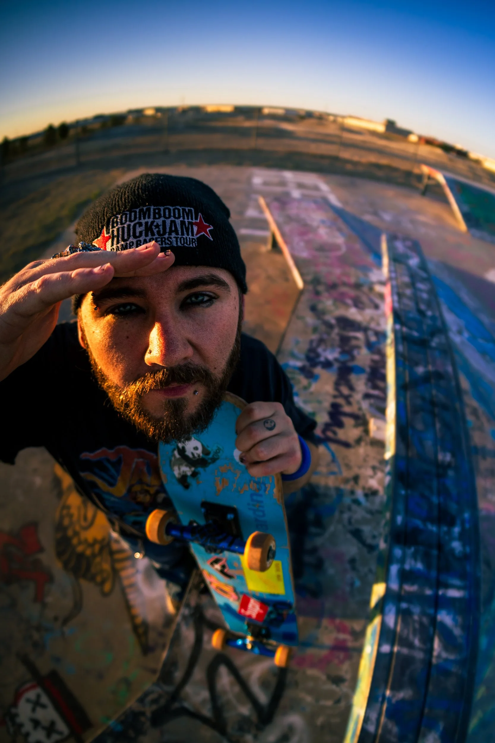 A young man looks into a fisheye lens as he holds a skateboard to his chest in a graffiti filled skatepark.