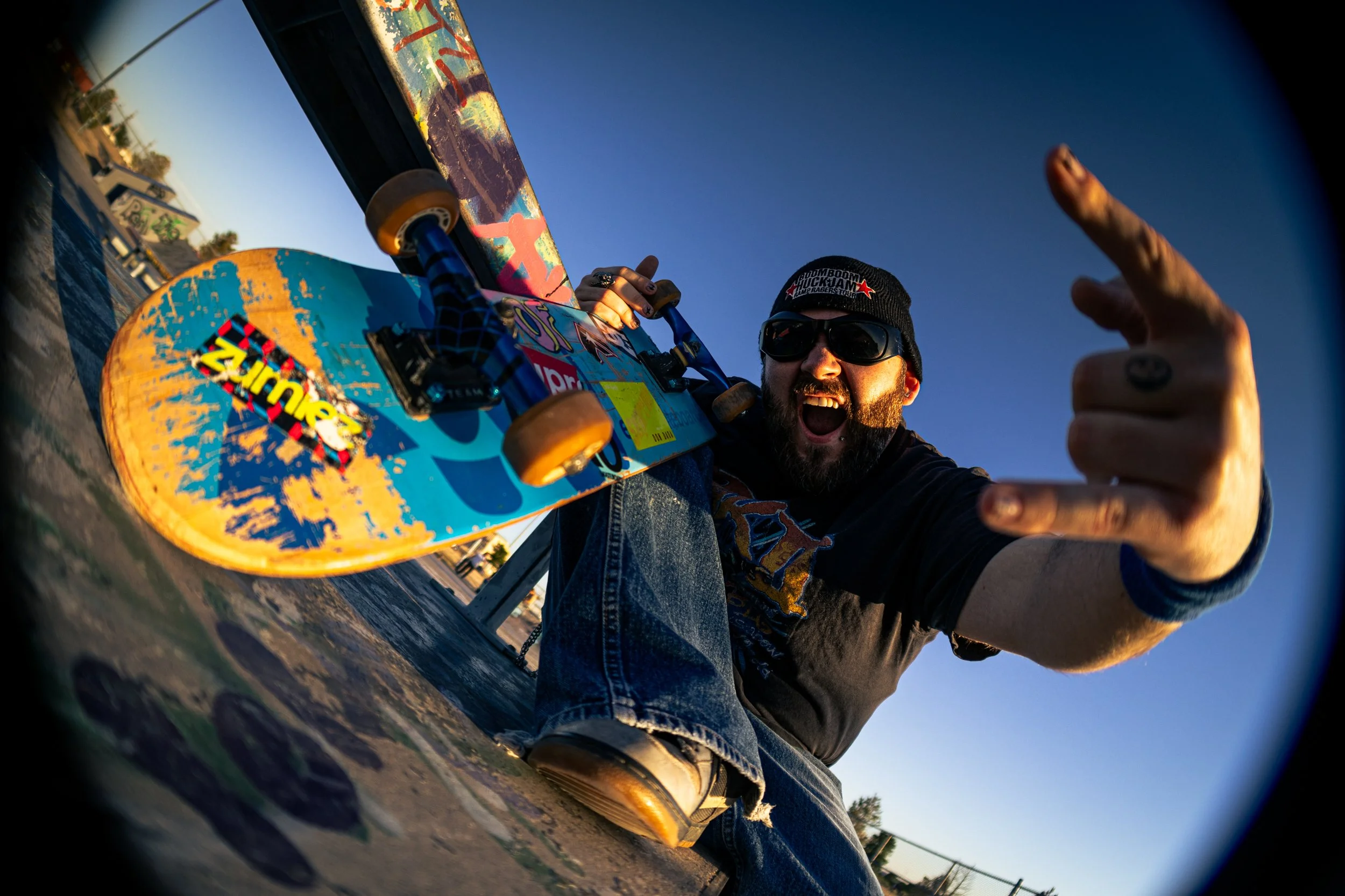 A young man in a beanie and sunglasses holds a skateboard in one hand and makes a rock sign with his other hand in a fisheye shot.