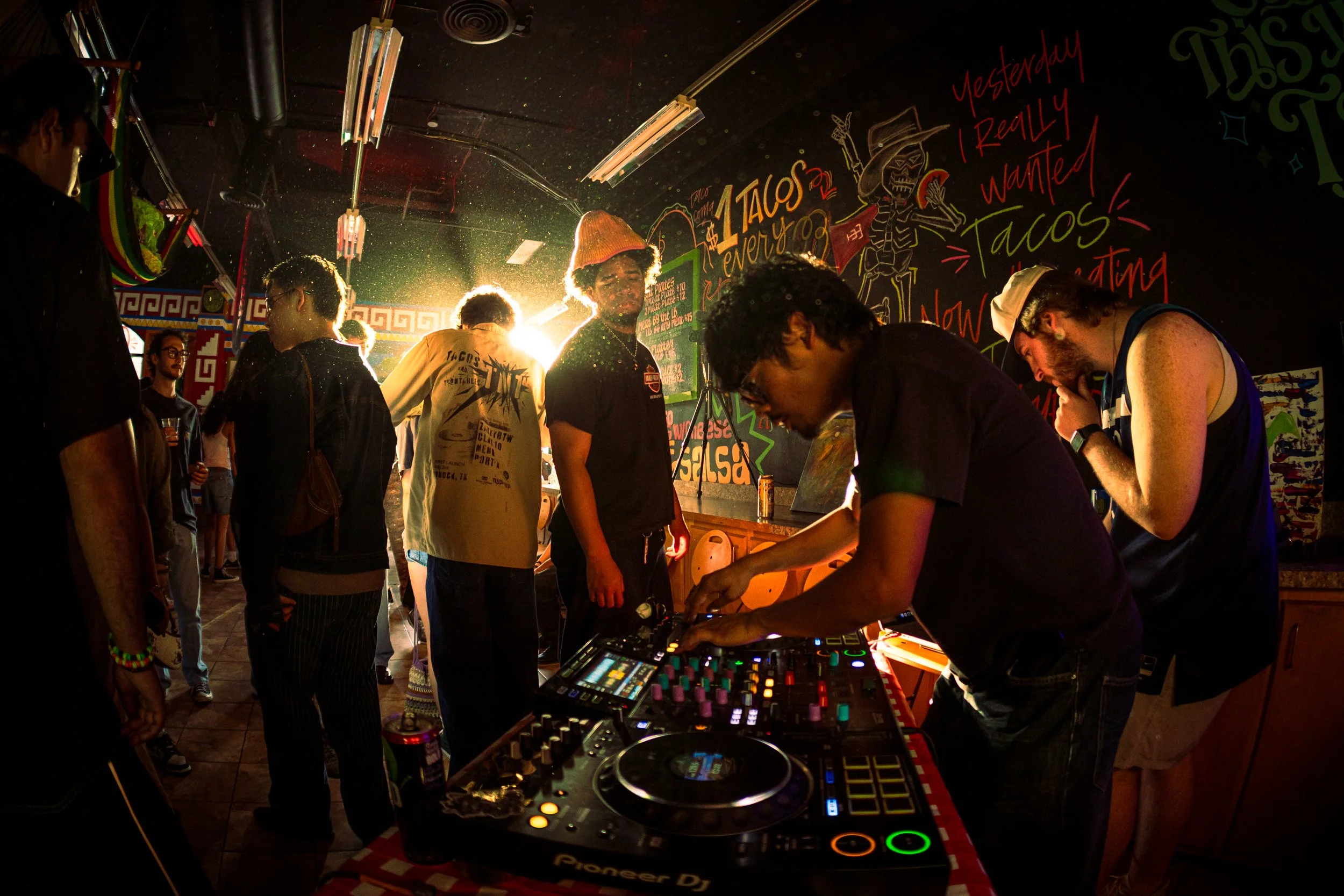 A live DJ mixing music in a small taco shop, causing patrons to get on their feet and dance. The lighting and graffiti filled background creates a colorful and energetic atmosphere.