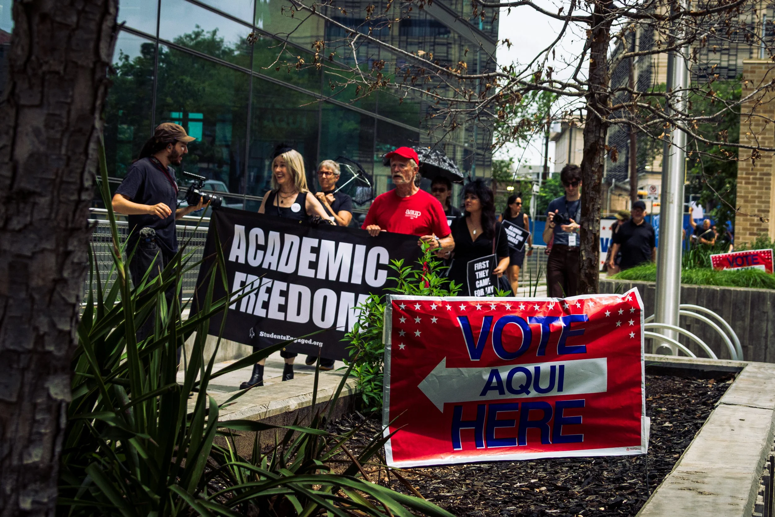 A red sign reading "Vote Aqui/Here" points toward a group of protestors marching while two of them hold a sign reading "Academic freedom" on a sidewalk next to downtown buildings .