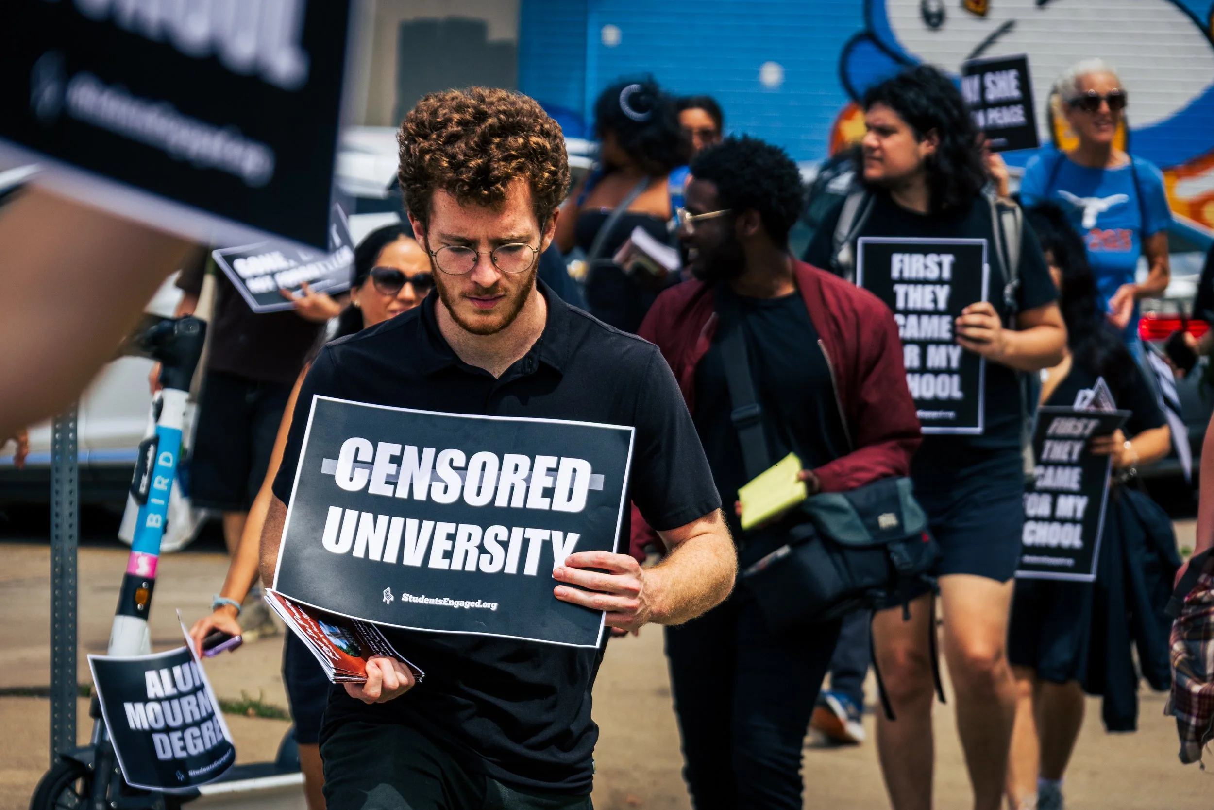 A group of college-aged youth hold signs, with the sign in focus reading "Censored University" as they march down an urban area.