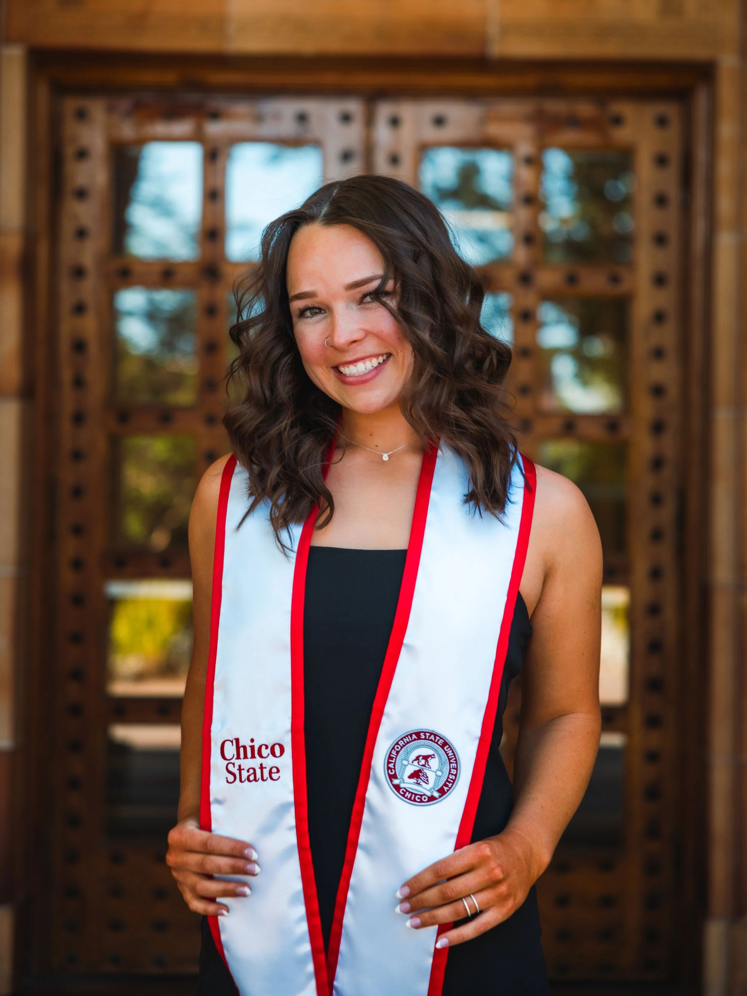 A smiling woman with wavy brown hair wearing a black top and a white and red Chico State stole standing in front of a wooden door with glass windows.