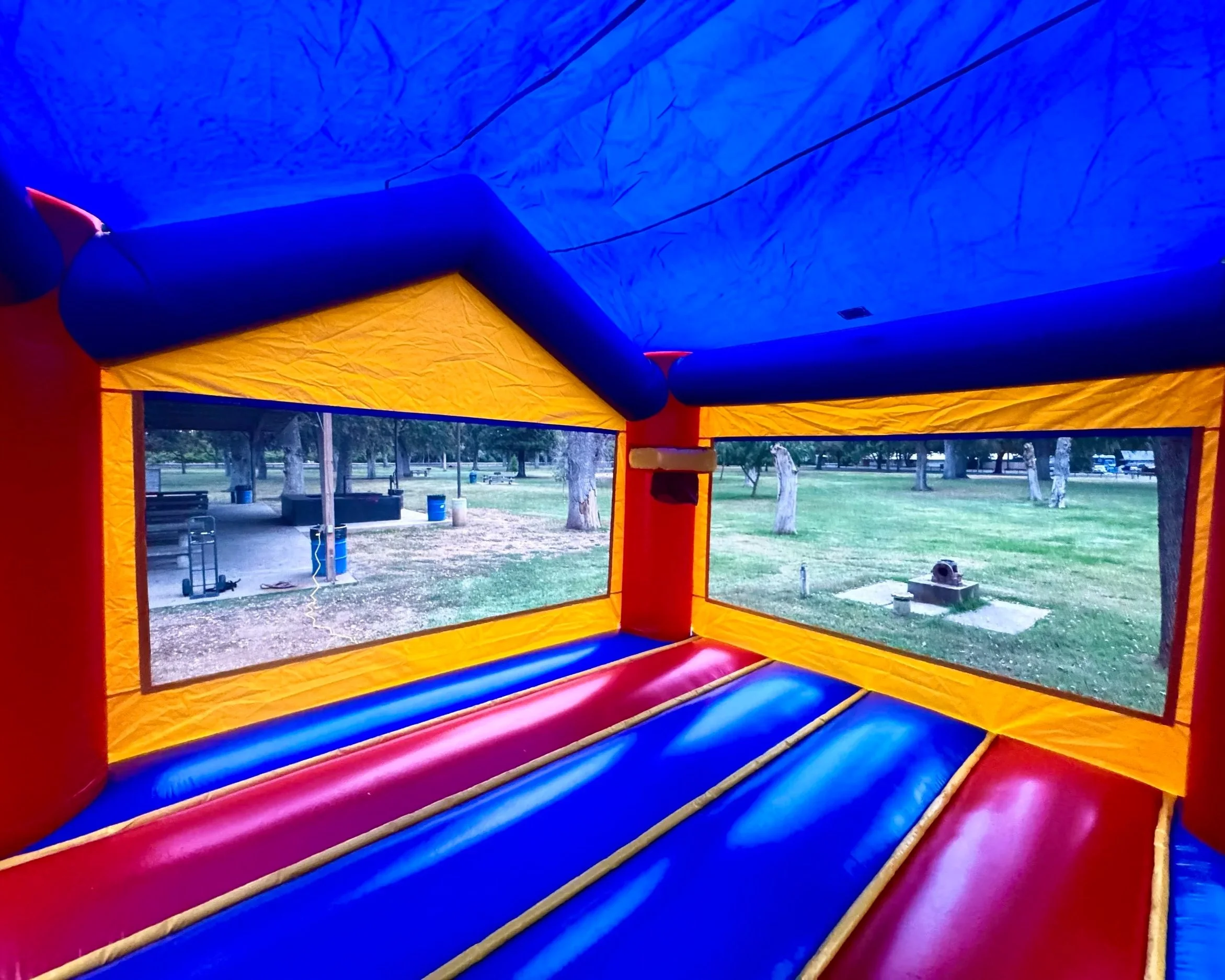 Inside view of a colorful inflatable bounce house with blue, yellow, and red sections, overlooking a park with trees and picnic tables outside.
