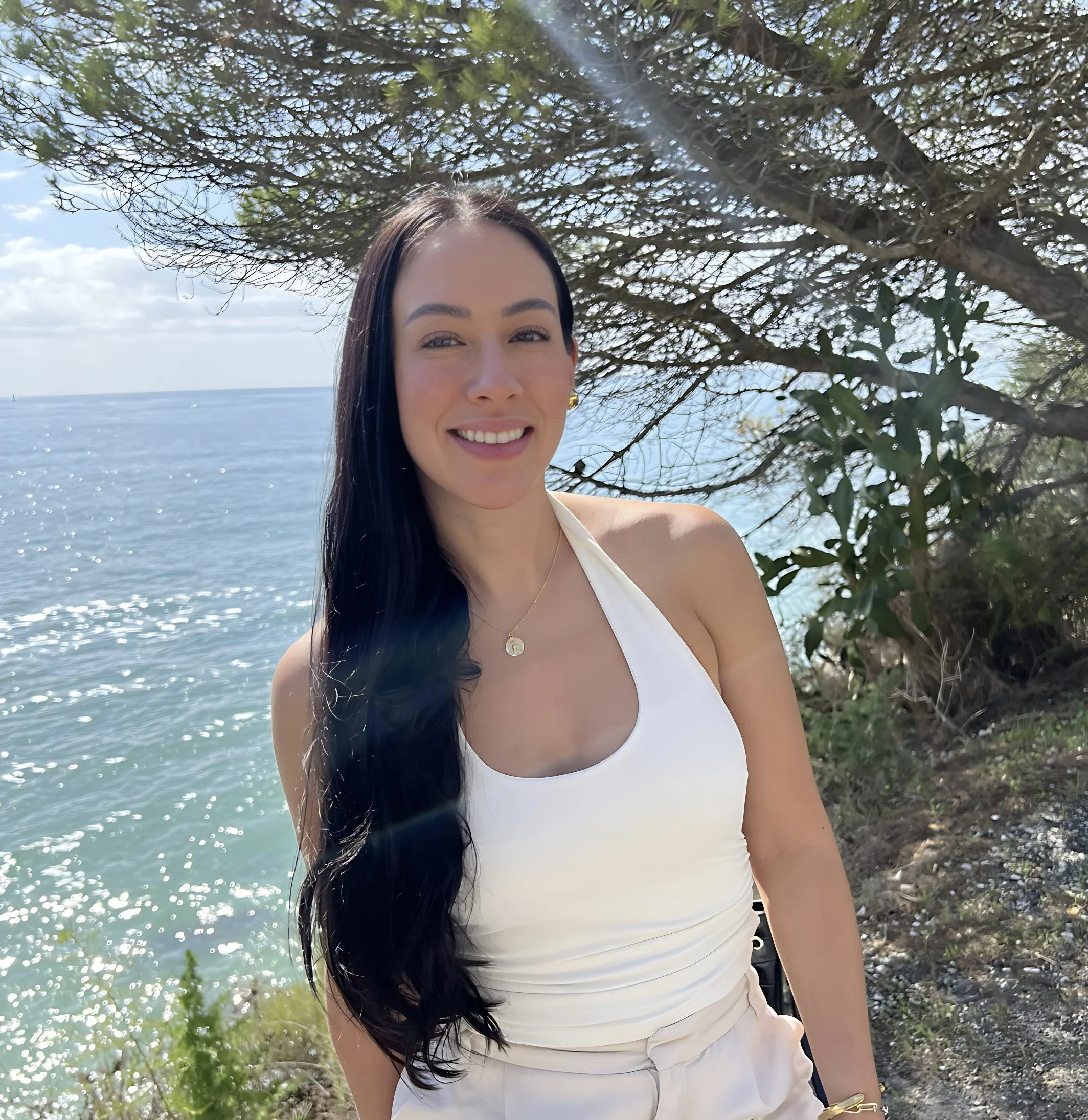 A woman with long dark hair smiling outdoors near the ocean, standing under a tree with green leaves