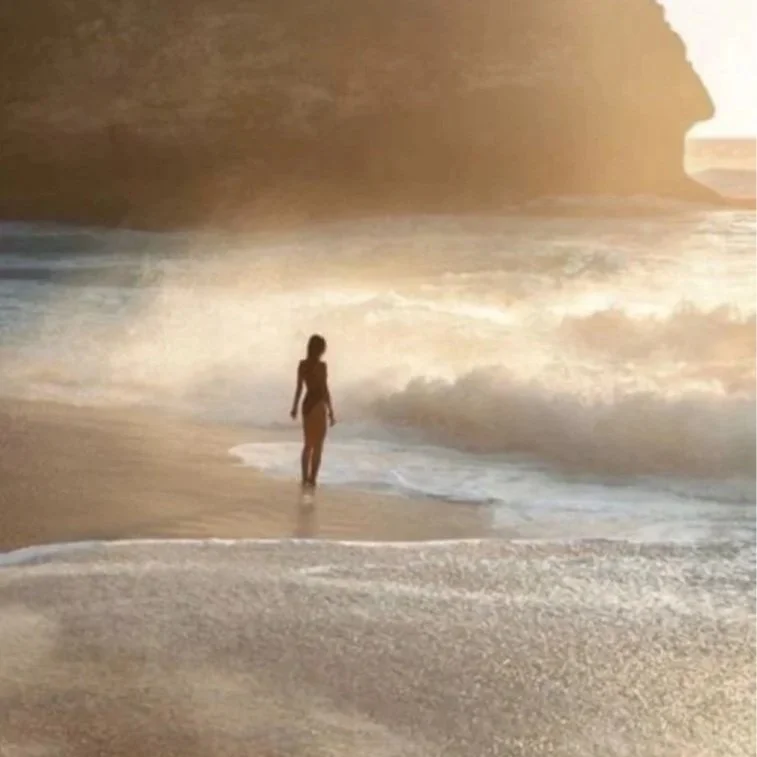 A woman walking along the shoreline on a beach during sunset with a rocky cliff in the background.