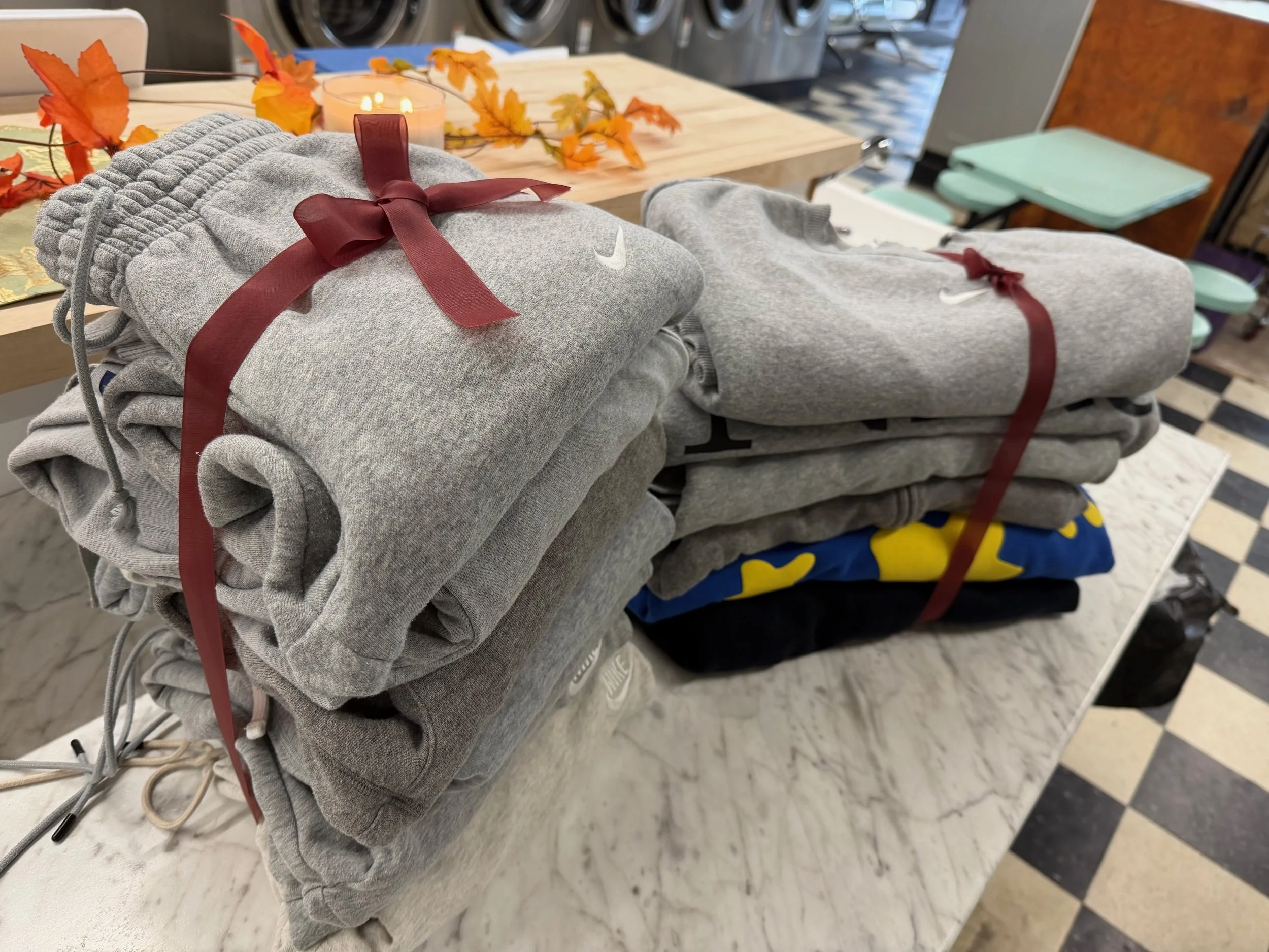 Stacks of folded sweatpants and hoodies tied with red ribbons on a marble table, with autumn leaves and candles in the background.