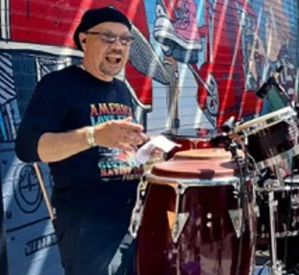 A man playing bongos while singing, wearing a black beanie and glasses, with a vibrant graffiti mural in the background.