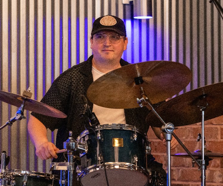 A man wearing a black cap with a cookie design, glasses, and a black shirt with white specks, sitting behind a drum set with cymbals, in a room with striped wall panels and blue lighting.