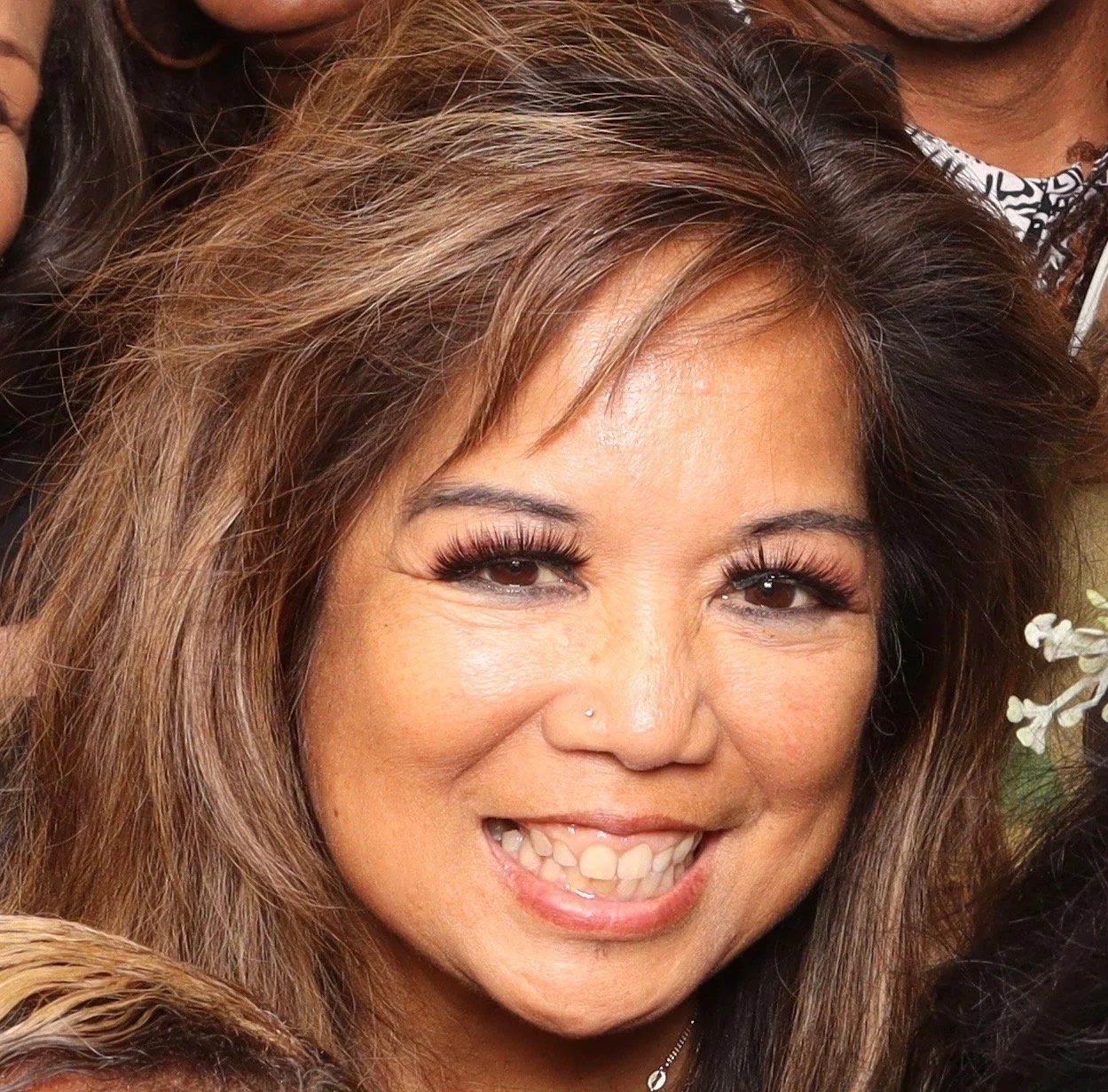 Close-up of a smiling woman with long, wavy brown hair, wearing makeup, a small nose piercing, and gold earrings, with a person partially visible in the background.