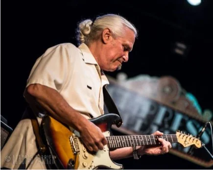 An elderly man with white hair playing an electric guitar on stage.