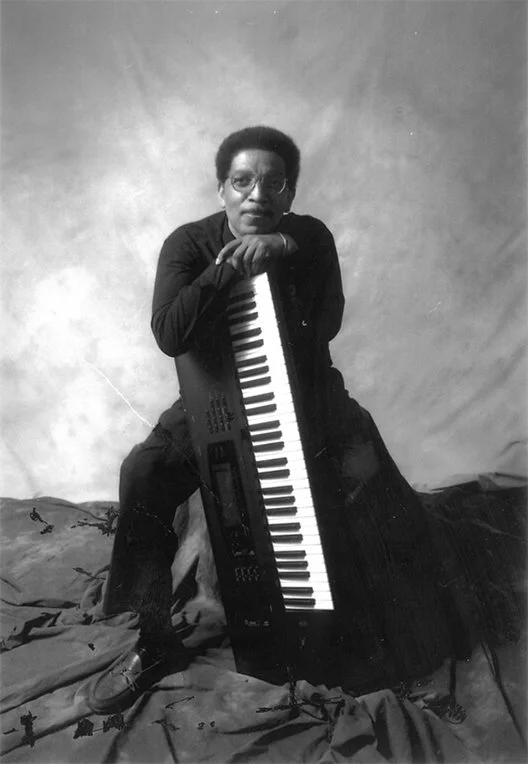 A person with short hair and glasses sitting on a stool, holding a keyboard instrument vertically in front of them, in a studio with a plain backdrop.
