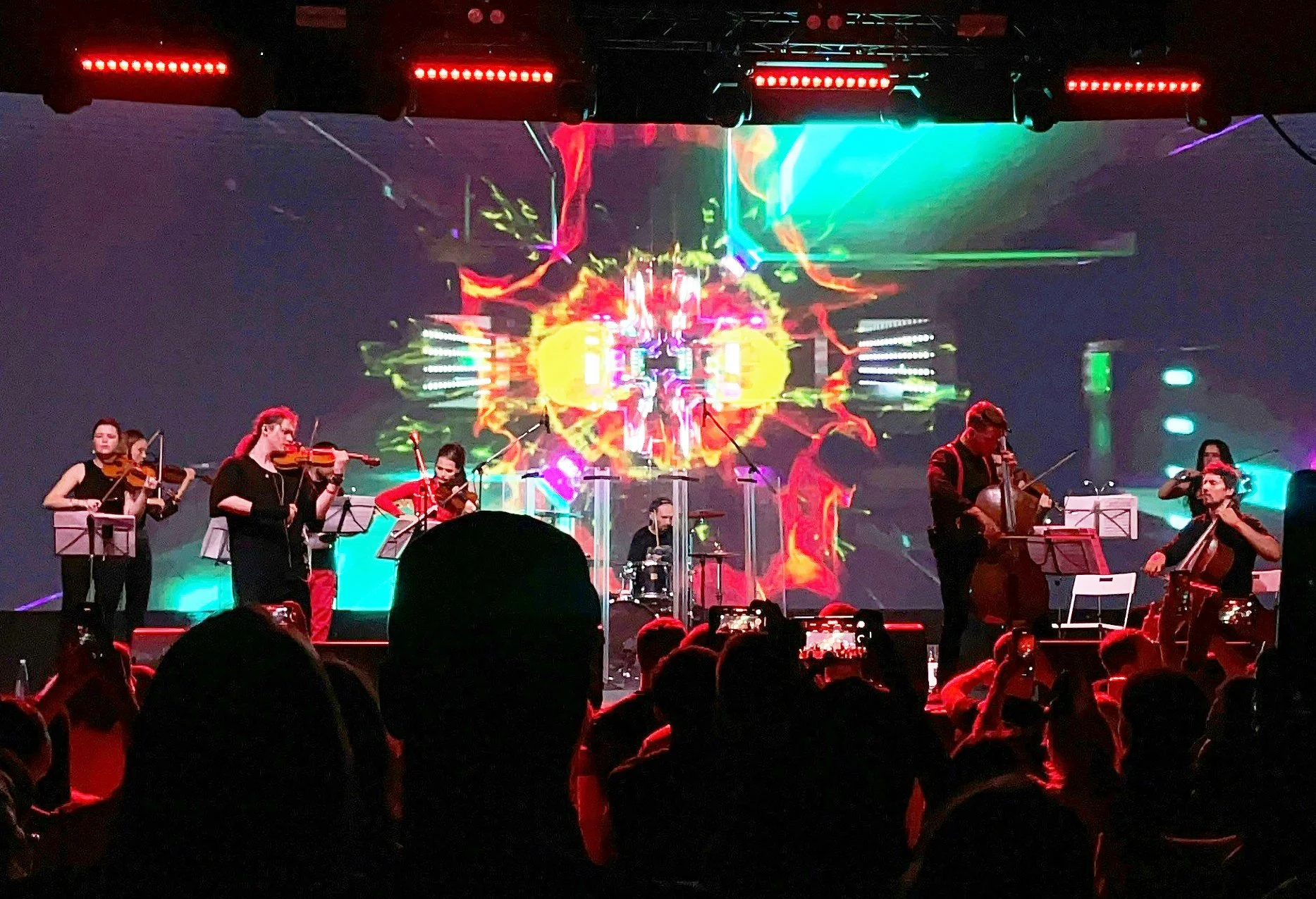 Stage performance with six musicians playing string instruments and drums, with colorful digital visuals on a large screen in the background and audience taking photos.