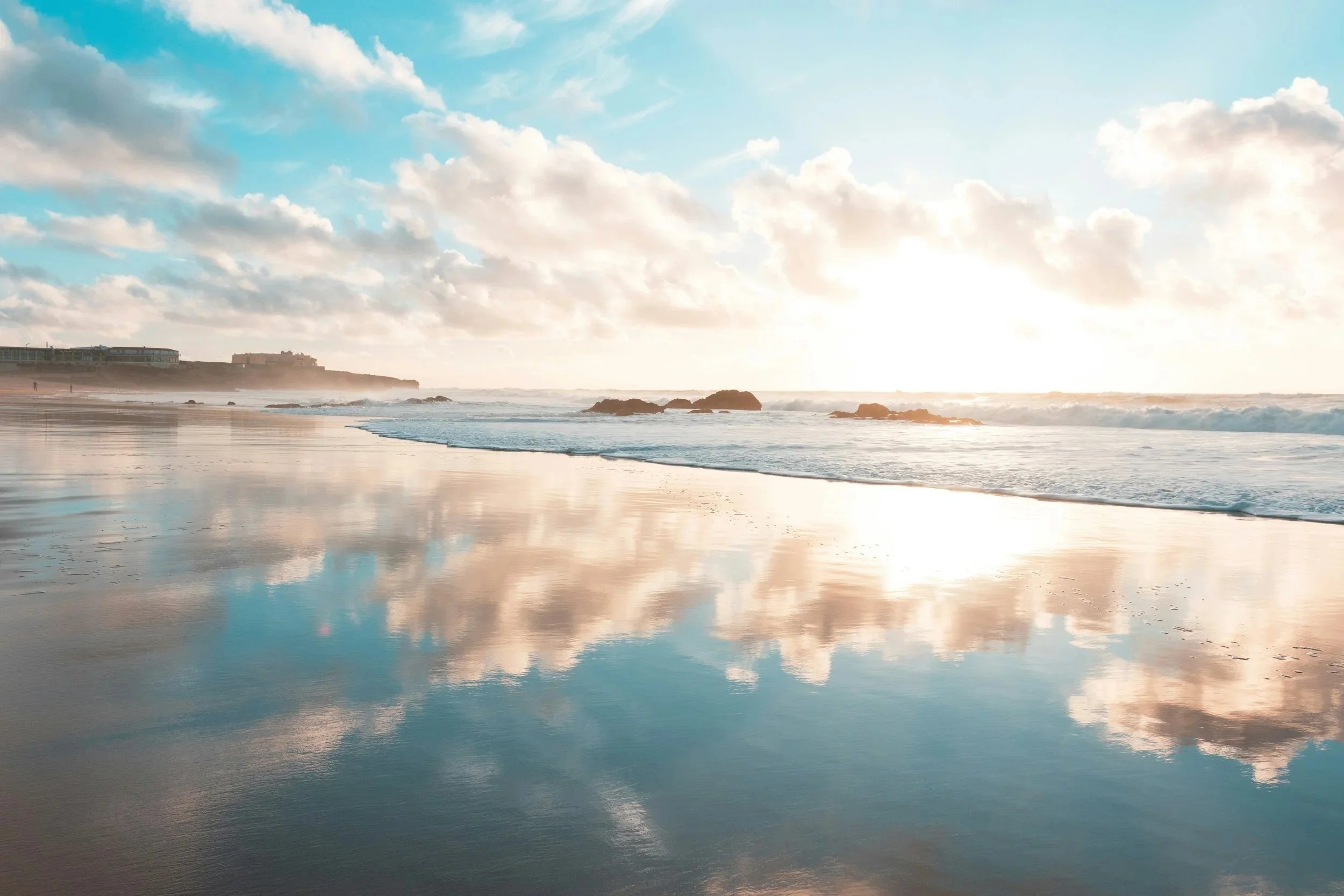 Sun partly visible over the ocean horizon with cloudy sky, reflecting on wet sandy beach, distant shoreline with rocks and buildings.