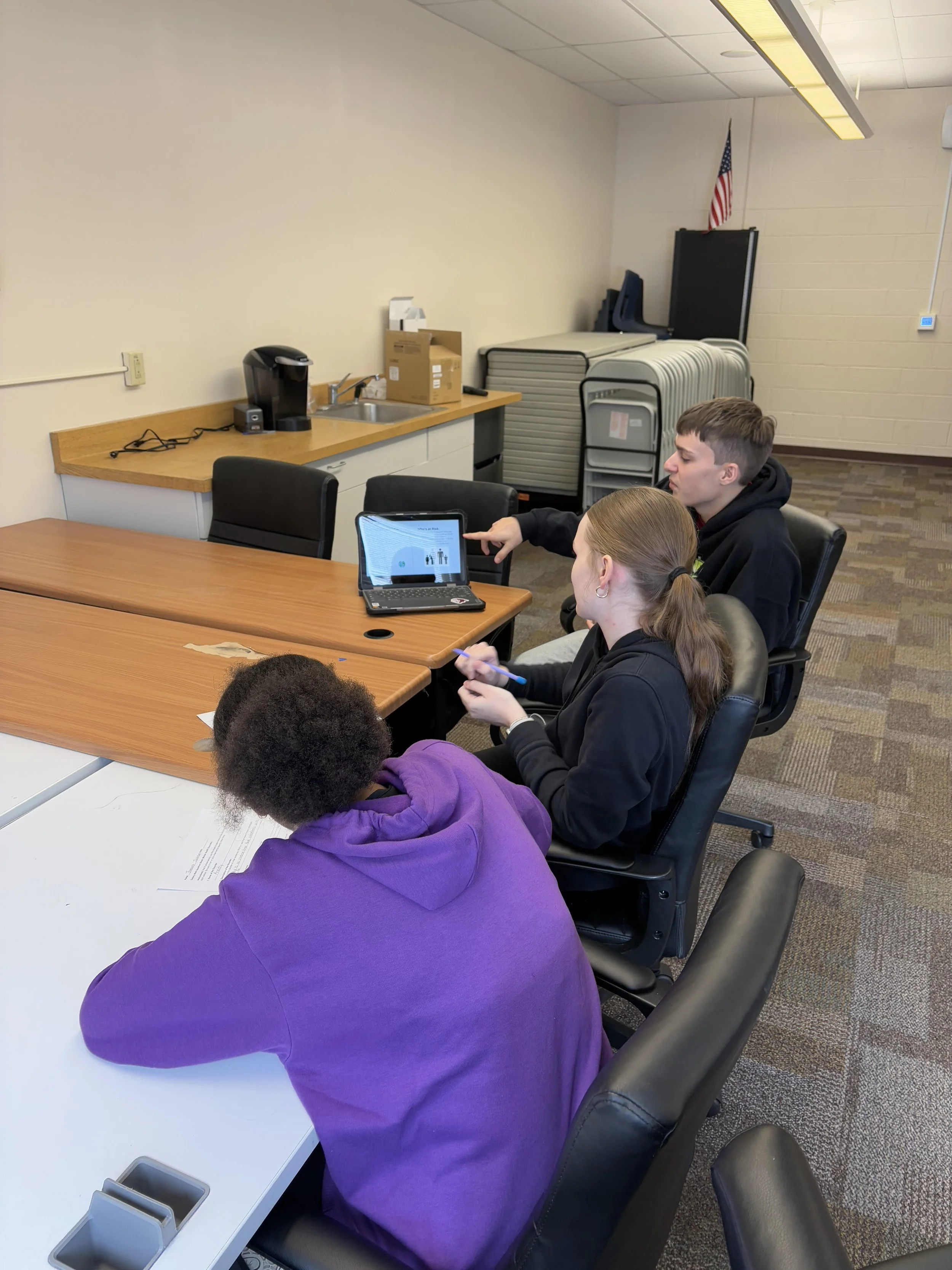 Three students sitting at a conference table looking at a laptop screen, with notes and a pen in hand, in a classroom or meeting room with a coffee station in the background.
