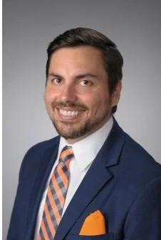 Professional headshot of a man with dark hair, beard, wearing a navy suit, white shirt, orange and navy striped tie, and orange pocket square, smiling against a gray background.