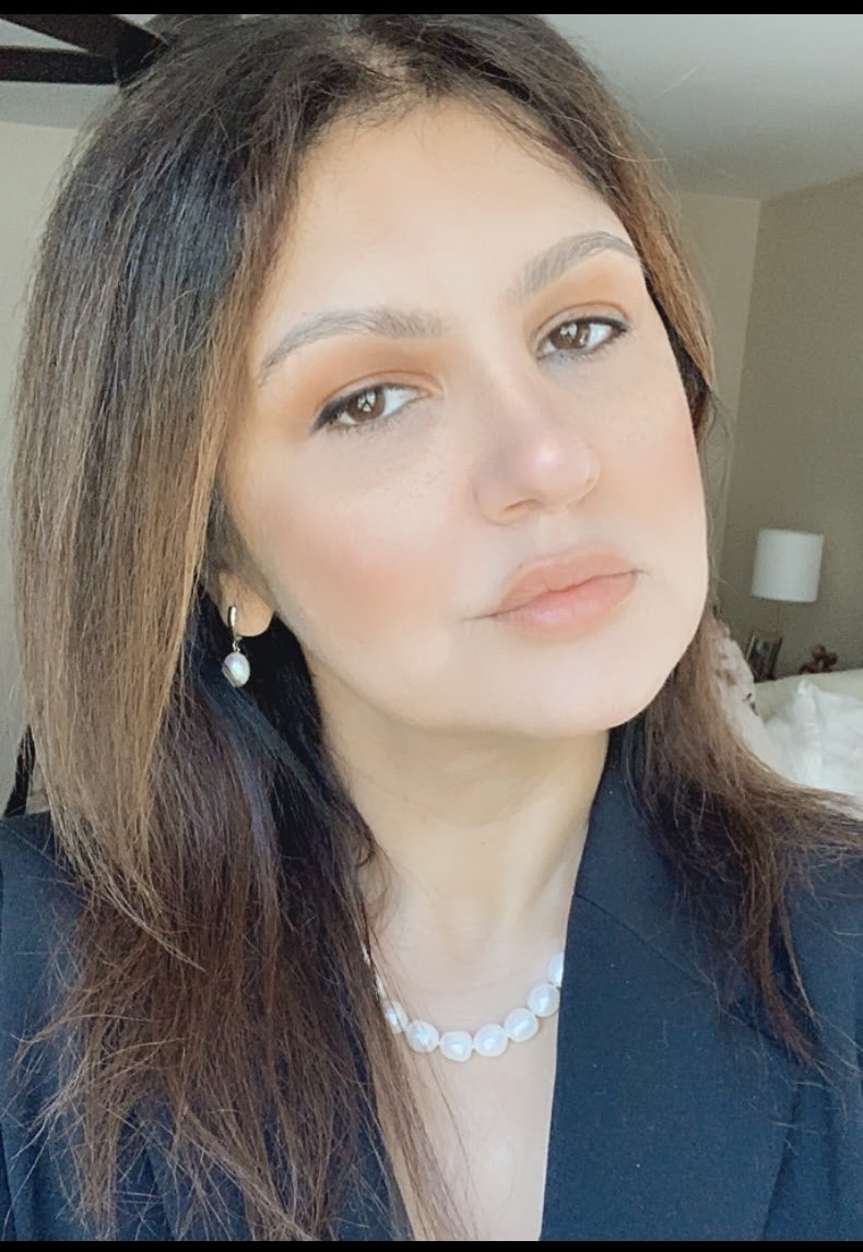Close-up of a woman with shoulder-length brown hair, wearing pearl earrings and a pearl necklace, dressed in a black top or blazer, in a room with a bed and lamp in the background.